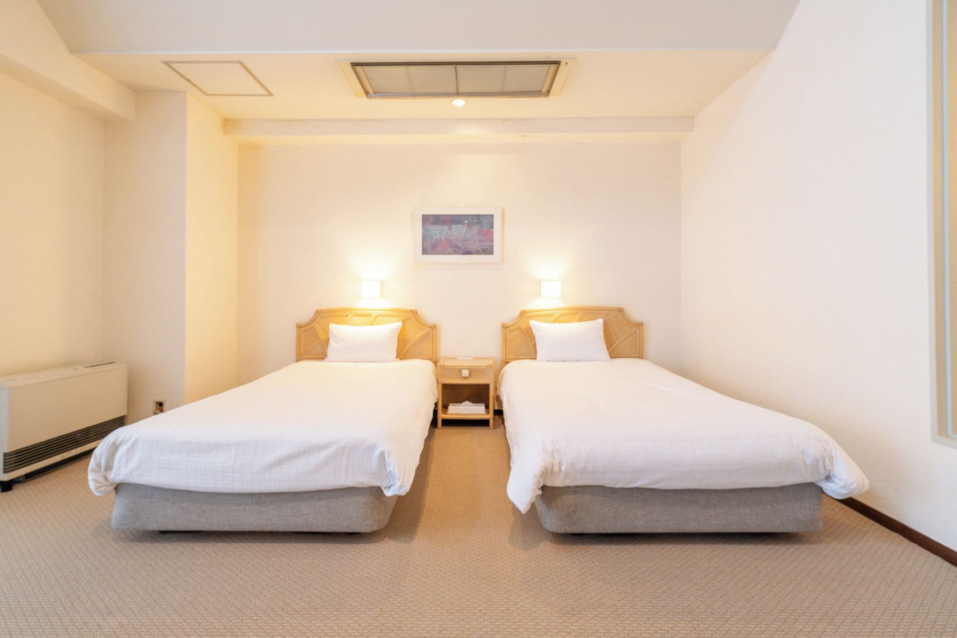 A hotel room with two single beds, a small bedside table with a lamp, a radiator, and a ceiling light fixture.