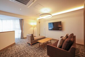 A hotel room with a sofa, a small table, a TV mounted on the wall, and a window with curtains. - Fuji Premium Resort (Fujikawaguchiko)