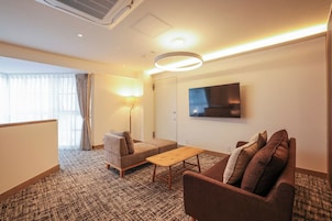 A hotel room with a sofa, a small table, a TV mounted on the wall, and a window with curtains.