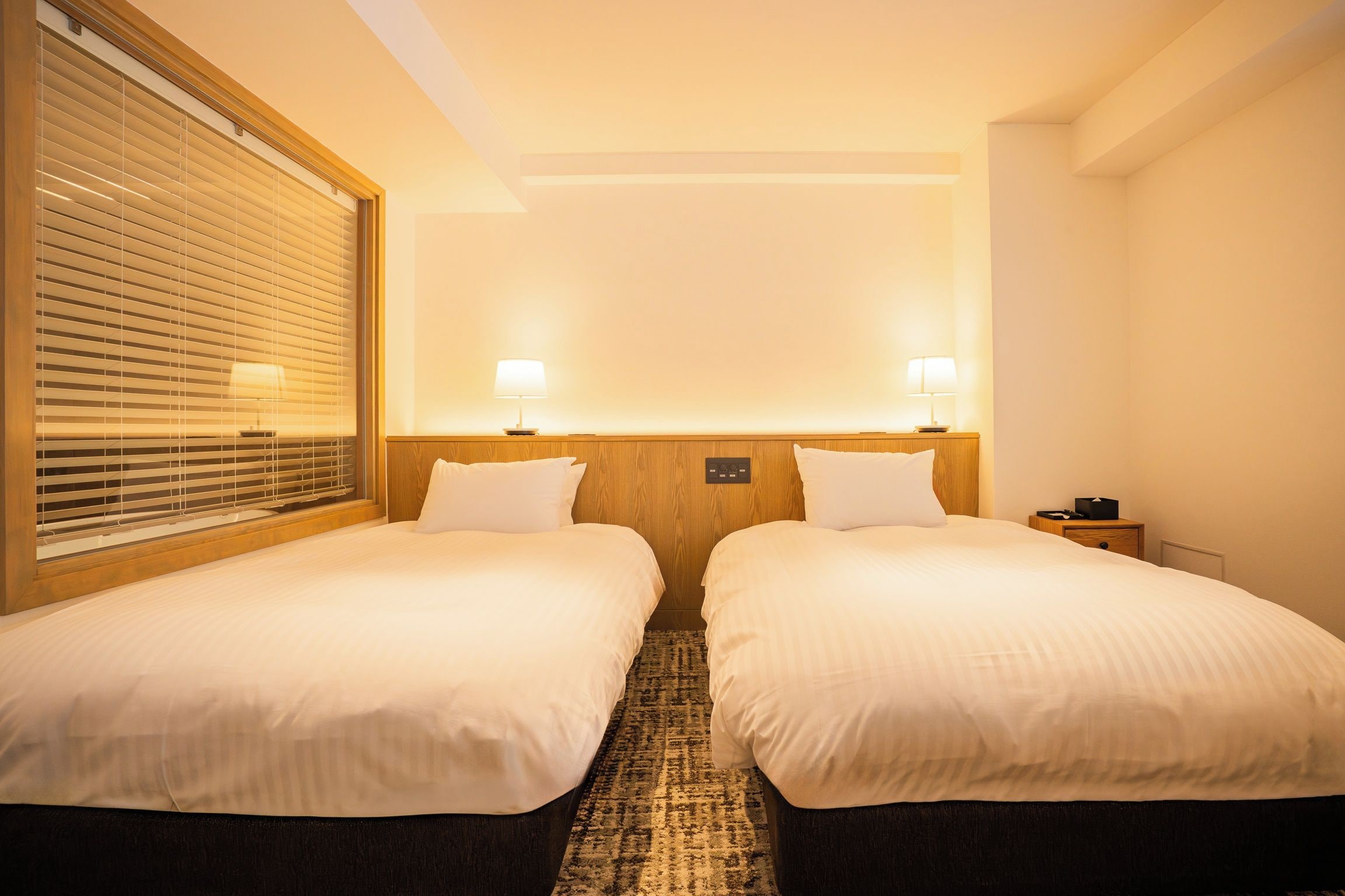 Una habitación de hotel con dos camas, una ventana con persianas, una mesita de noche con lámpara y suelo alfombrado.