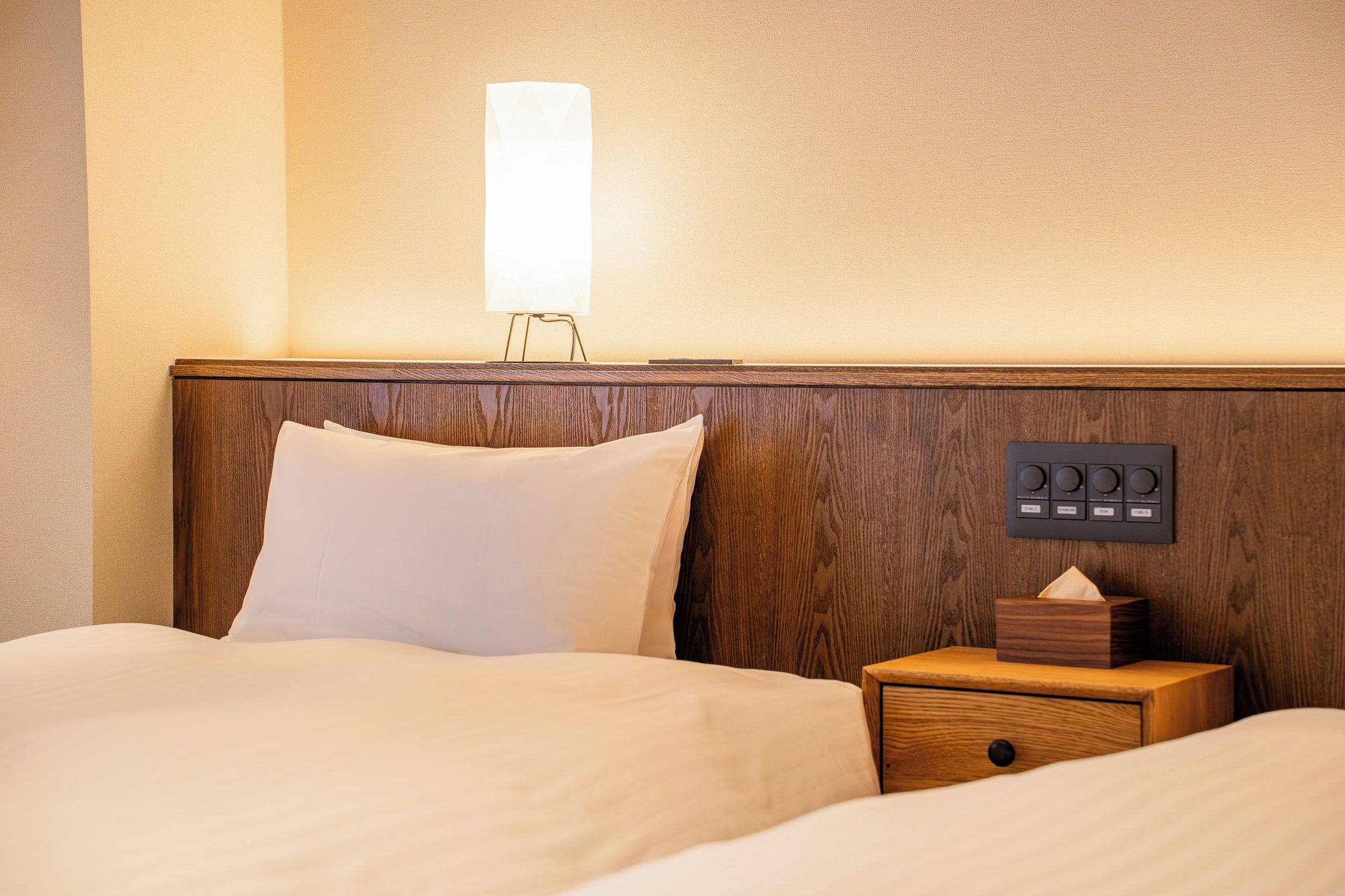 A hotel room with a wooden headboard, a bedside table with a tissue box, and a wall-mounted control panel.
