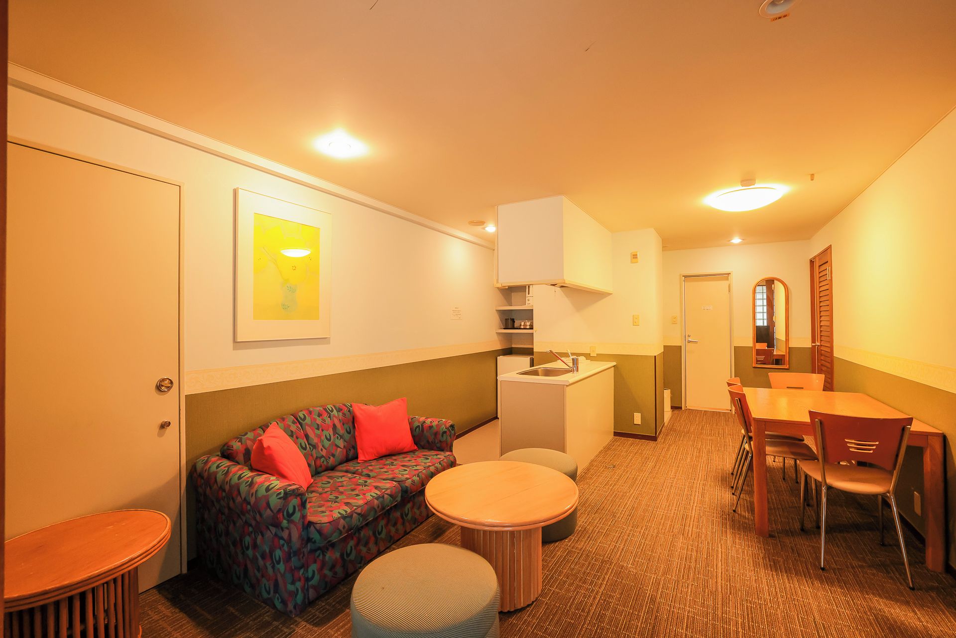 A compact hotel room with a sofa, a small dining table, a kitchenette, and a mirror.