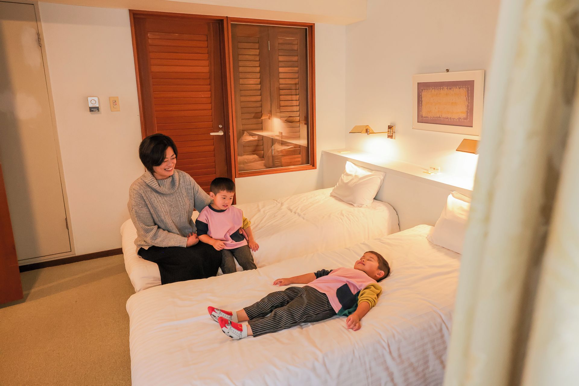 A hotel room with a bed, a bedside table with a lamp, a window with blinds, and a door with a lock.