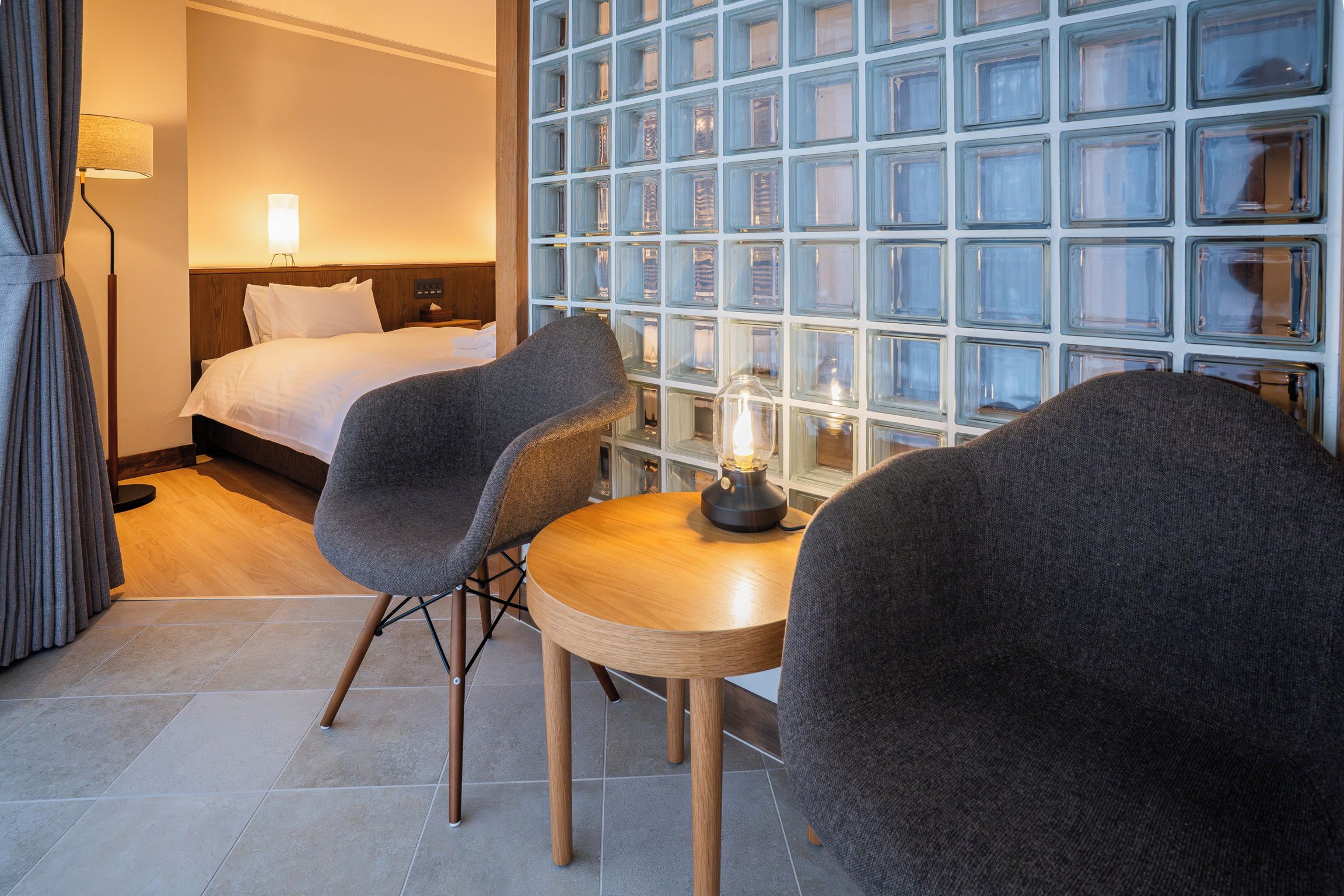 A hotel room with a bed, two armchairs, a small wooden table, and a wall of glass blocks.