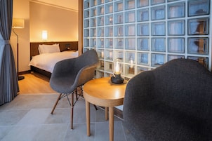 Habitación de hotel con una cama, dos sillones, una mesita de madera y una pared de bloques de vidrio.