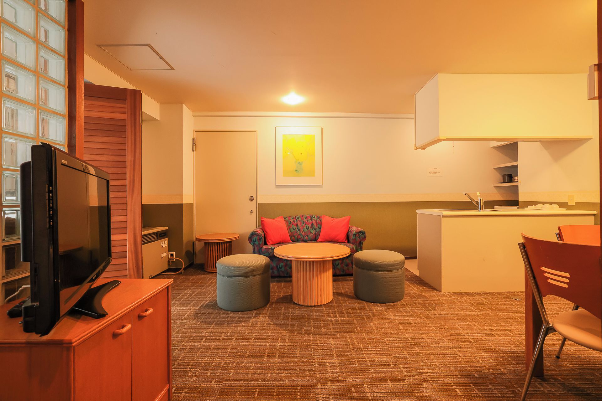A hotel room with a television, a sofa, two ottomans, a small table, and a kitchenette area.