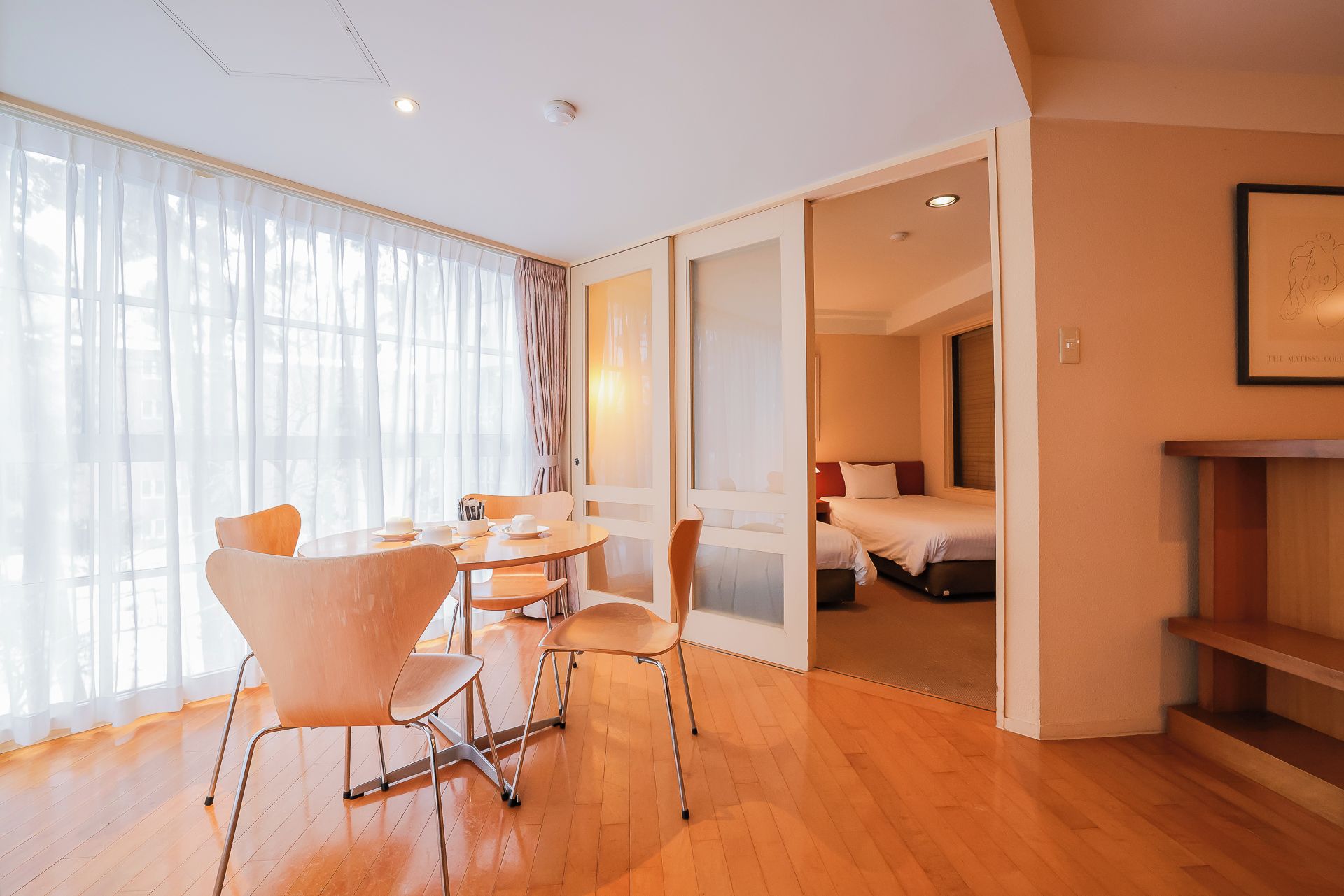 A hotel room with a wooden floor, a dining area with chairs and a table, and a bed with a red headboard.