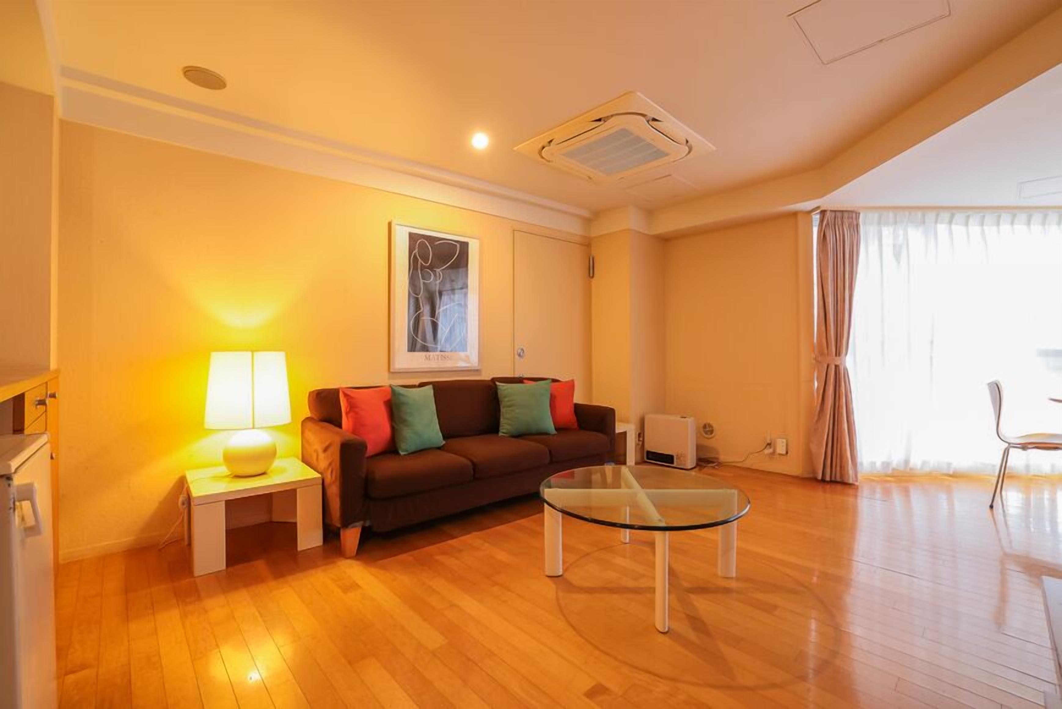 A living room with a sofa, a glass coffee table, a lamp, a painting on the wall, and a window with curtains.