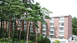 A multi-story building with a red facade and white windows, surrounded by a well-maintained lawn and trees. - Fuji Premium Resort (Fujikawaguchiko)