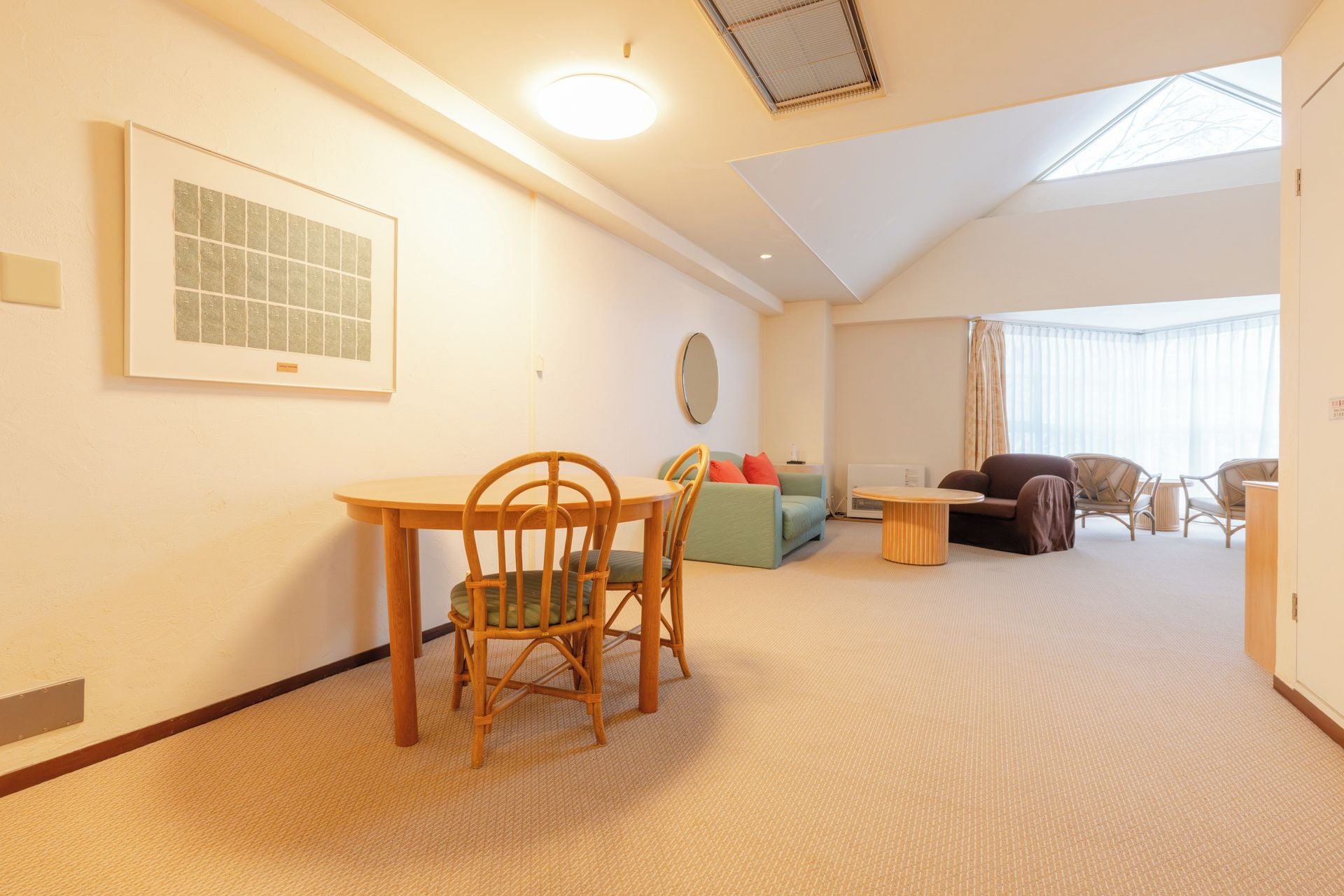 A spacious living area with a dining table, chairs, a sofa, and a large window providing natural light.