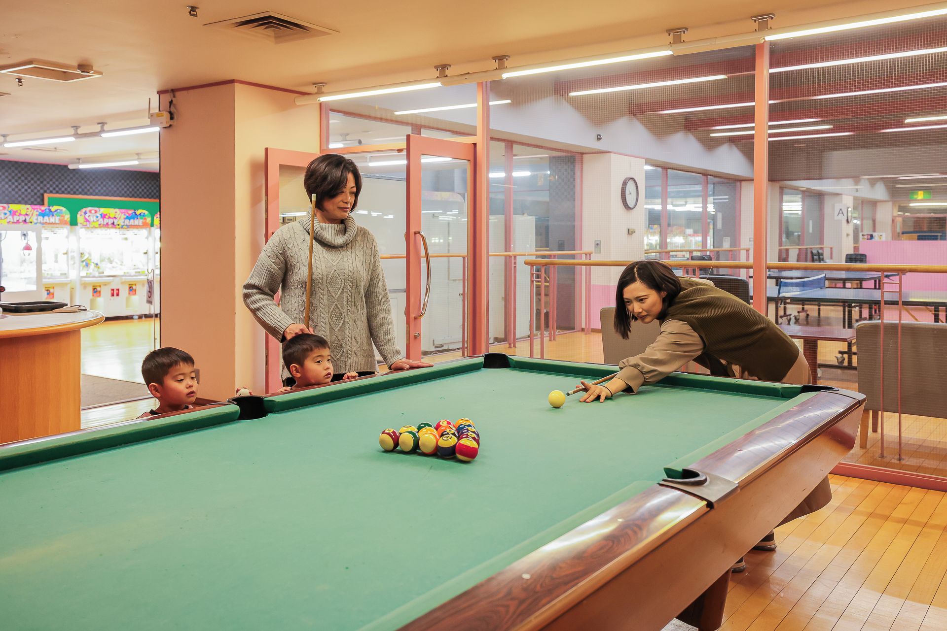 A pool table in a room with a wooden floor, a glass partition, and a clock on the wall.