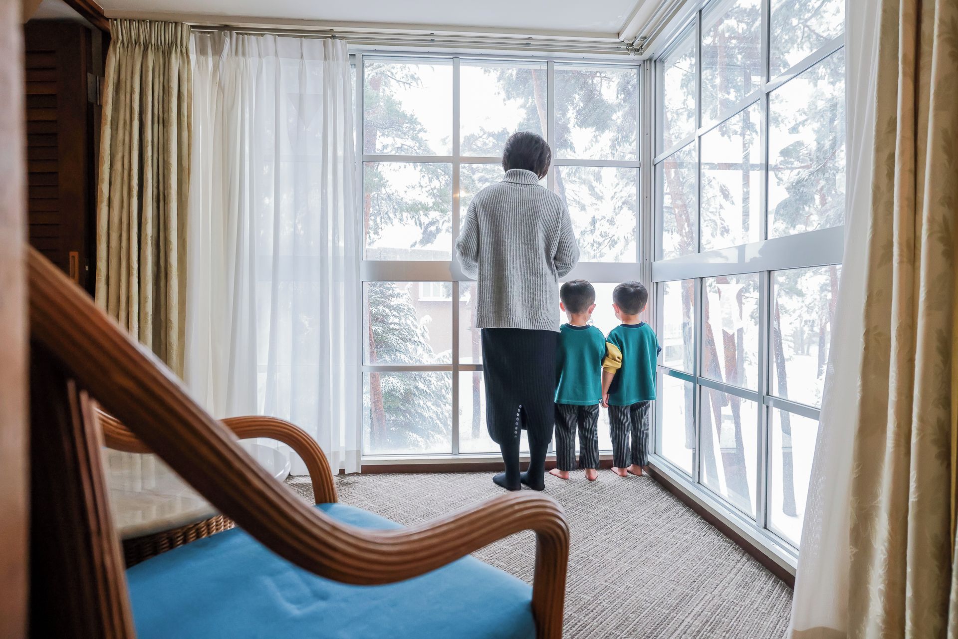 A room with a large window, a wooden chair, and a person standing with two children.