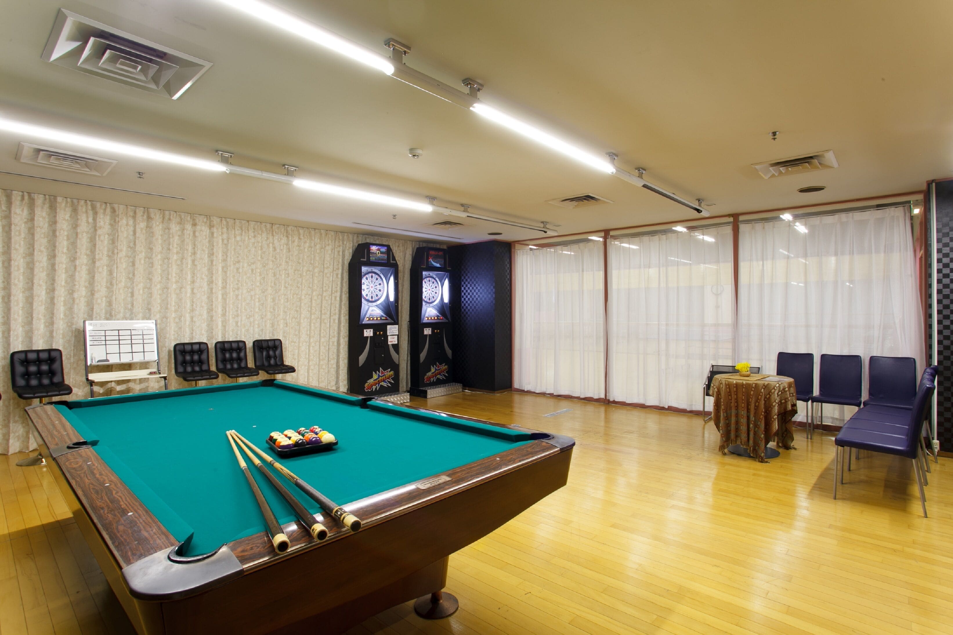 Una mesa de billar en una habitación con piso de madera, una mesa con sillas y un gran ventanal con cortinas.