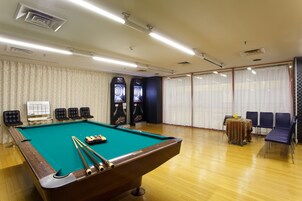 Una mesa de billar en una habitación con piso de madera, una mesa con sillas y un gran ventanal con cortinas.