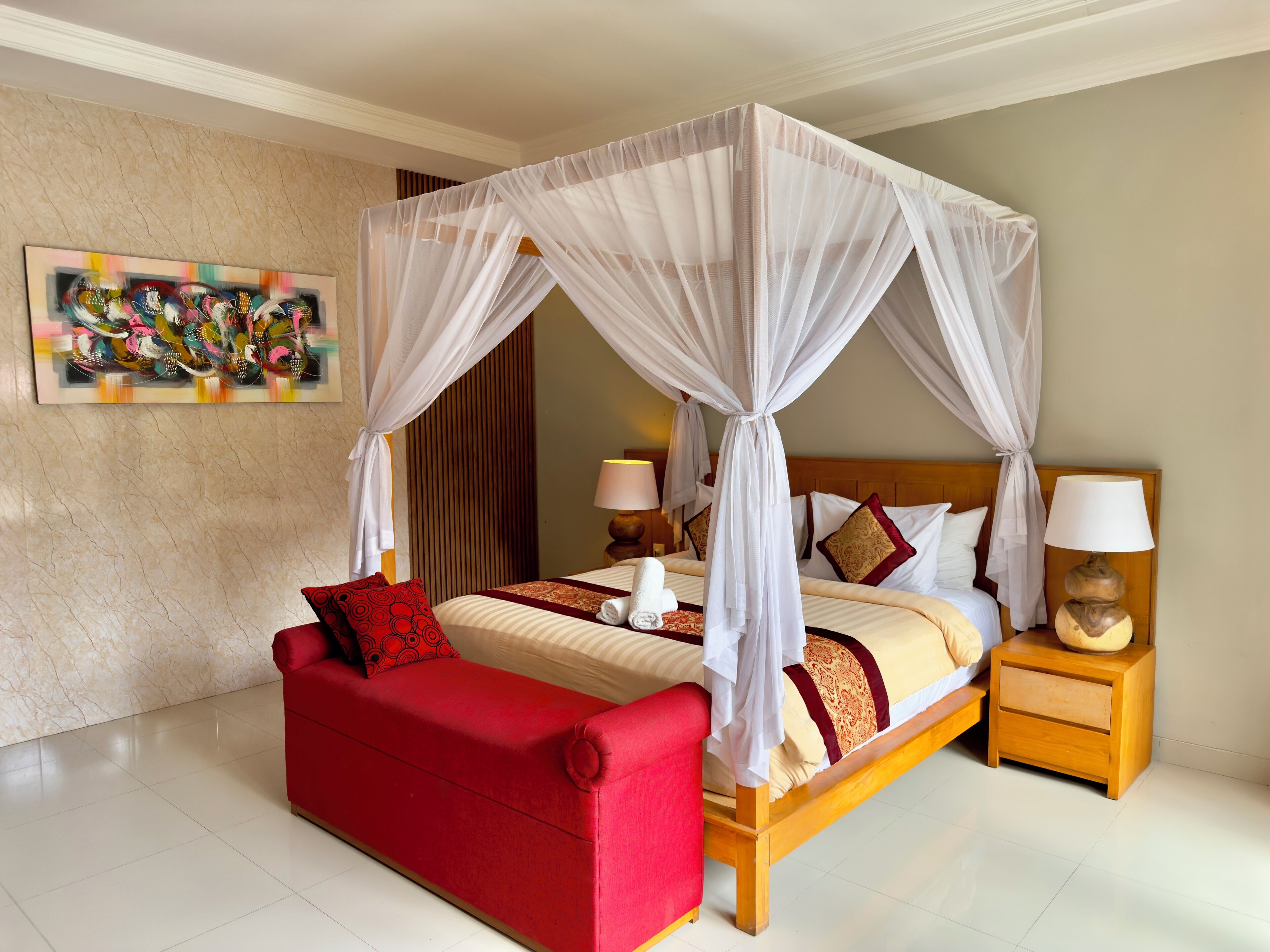 A four-poster canopy bed with a red sofa and wooden nightstands, a marble wall, and a colorful painting.