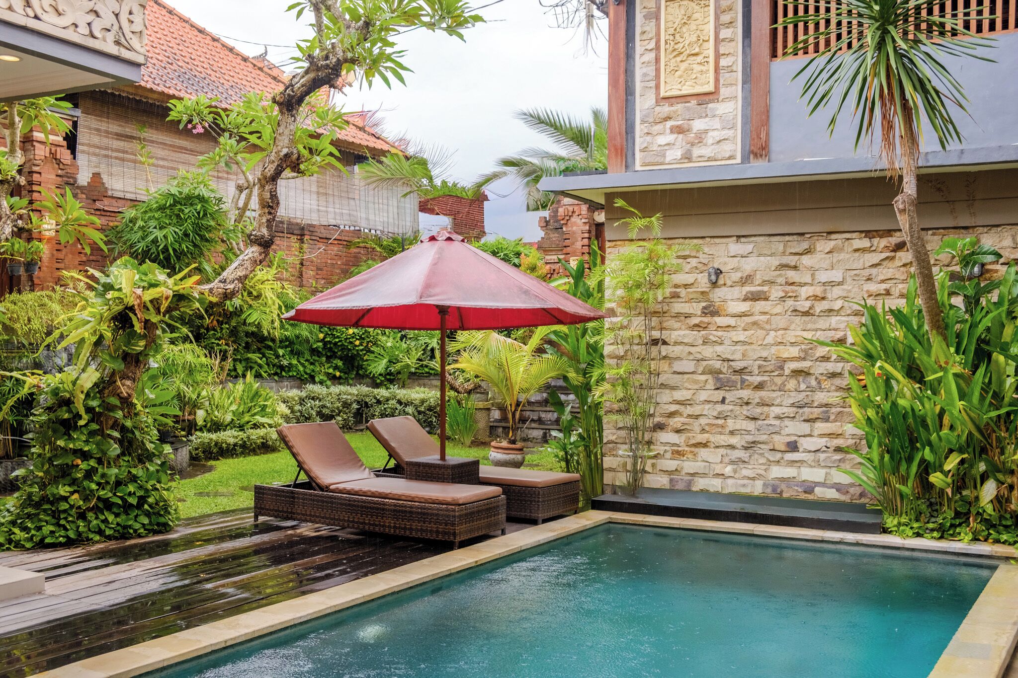 A pool area with lounge chairs and a red umbrella, surrounded by lush greenery and a stone wall.