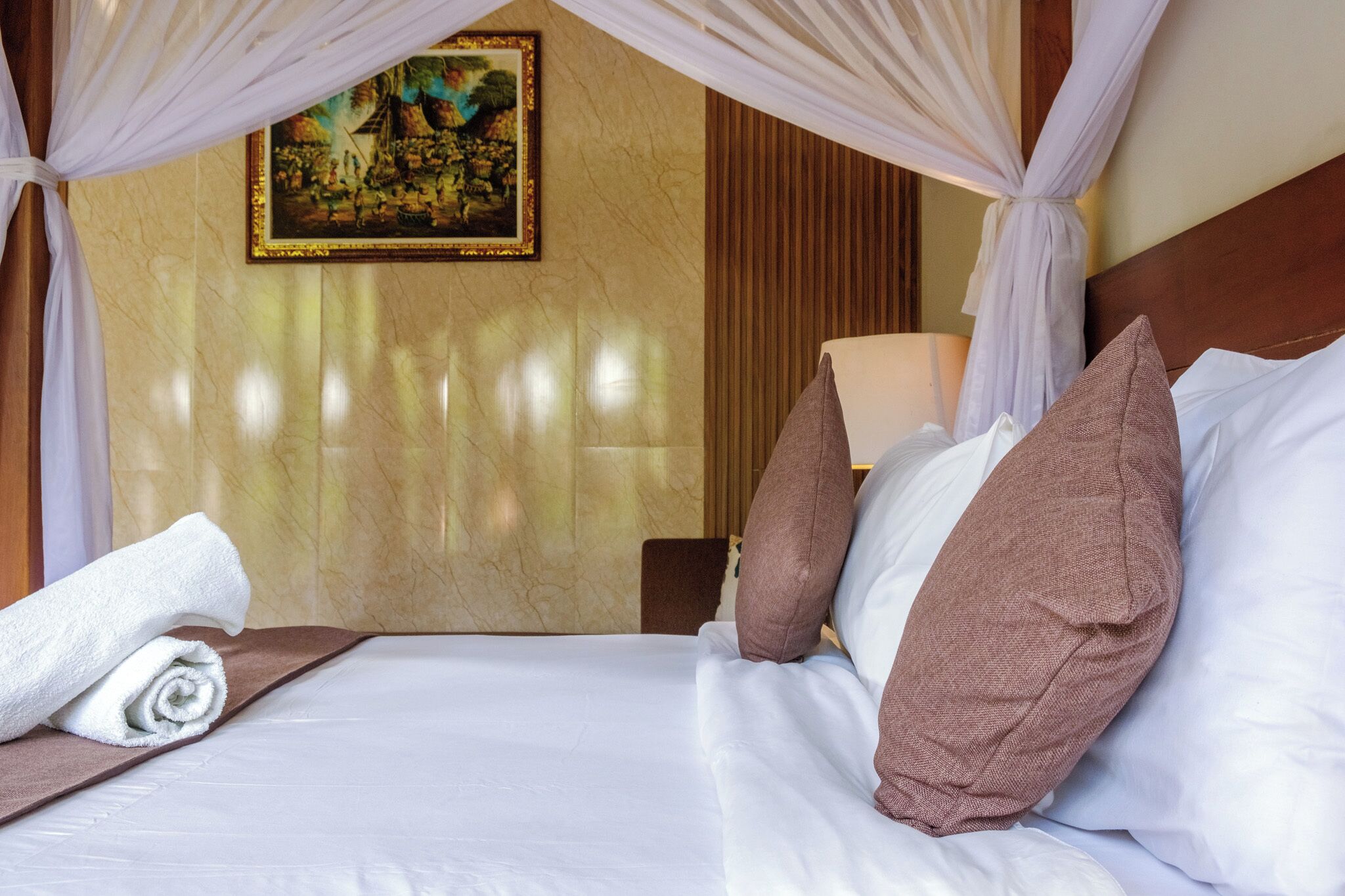 A hotel room with a bed, a rolled white towel, a painting on the wall, and a wooden headboard.