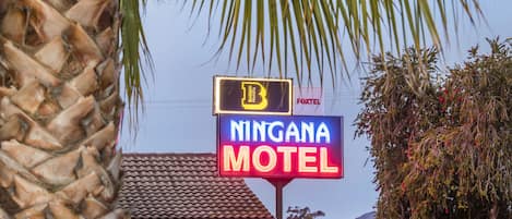 Señal de motel con los logotipos 'Ningana Motel' y 'Budget Motel', rodeada de palmeras y un cielo despejado.