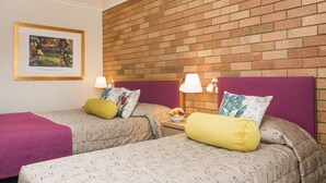 A hotel room with two beds, each with patterned bedding and pillows, a brick wall, a framed picture on the wall, and a small table with a lamp and fruit bowl. - Ningana Motel (Mudgee)