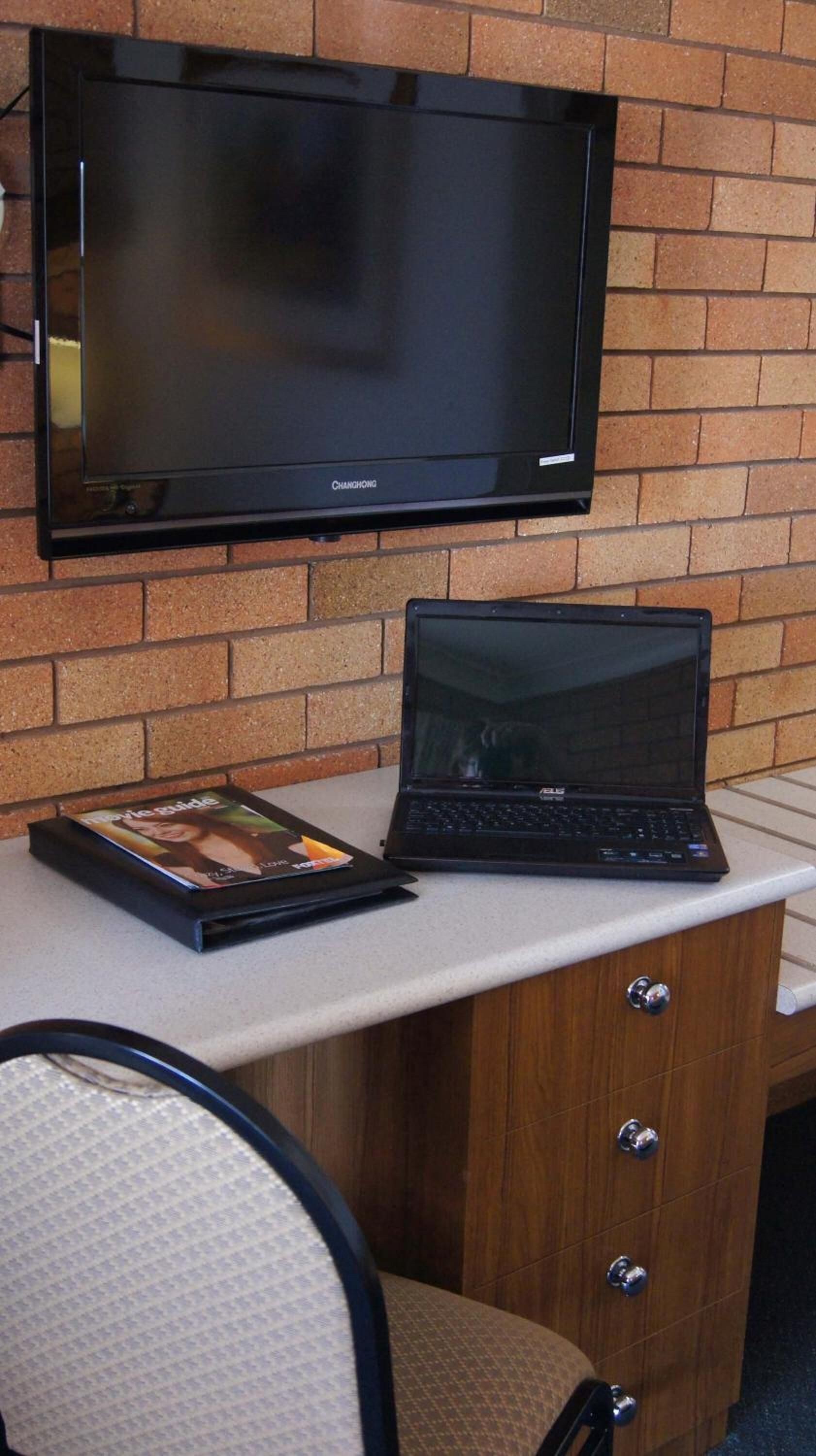 A flat-screen TV mounted on a brick wall, a laptop on a desk, and a magazine titled "Movie Guide".