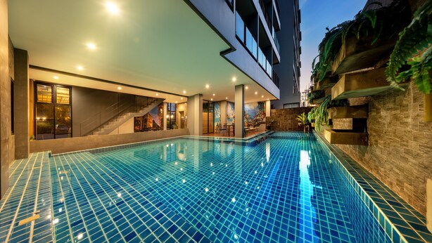 A modern indoor swimming pool with a tiled floor, surrounded by a building with large windows and a staircase. - The Wind (Sattahip)