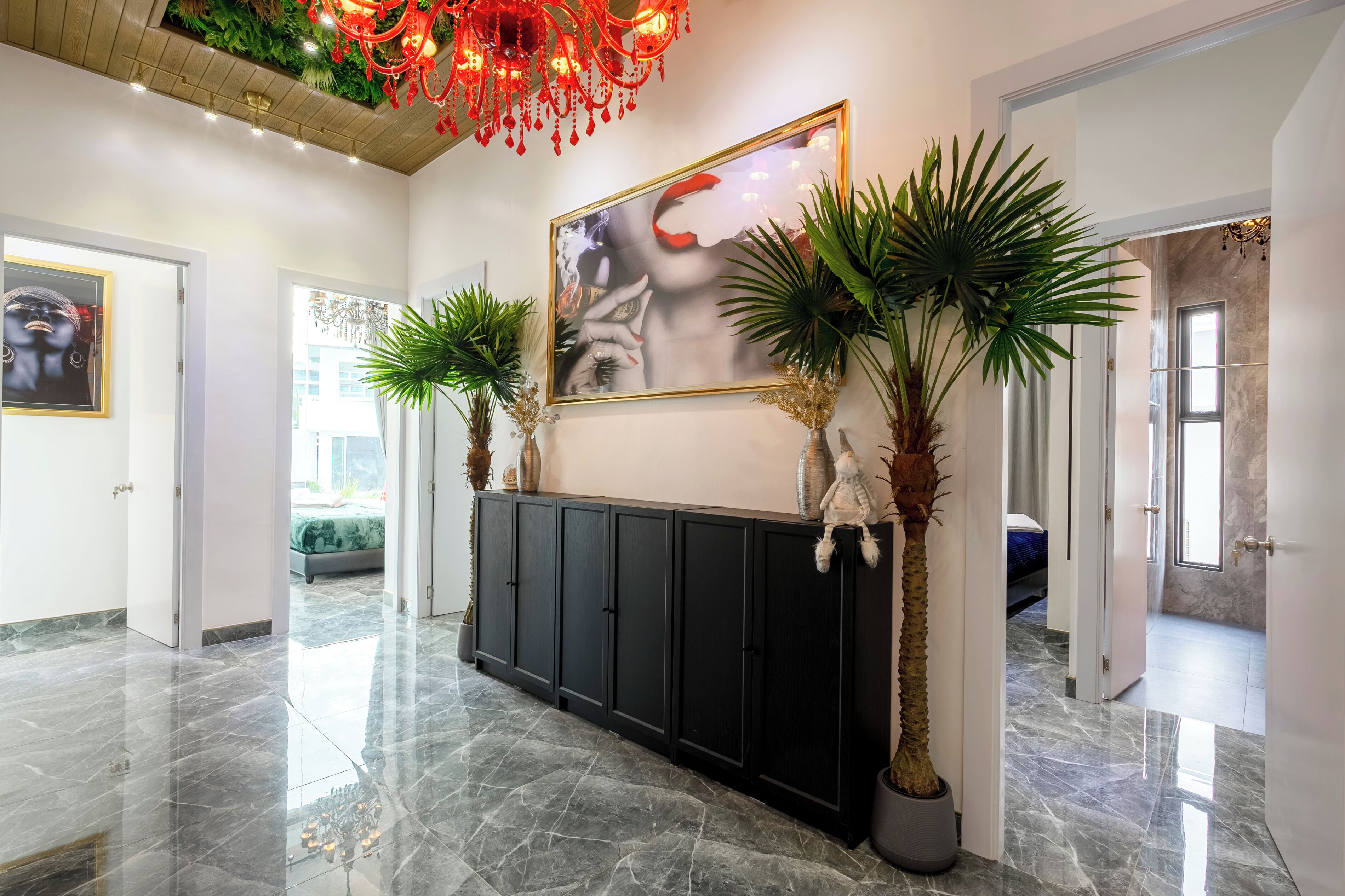 Een moderne lobby met een marmeren vloer, een zwarte console tafel met decoratieve planten, een groot kunstwerk in een lijst en een rode kroonluchter.