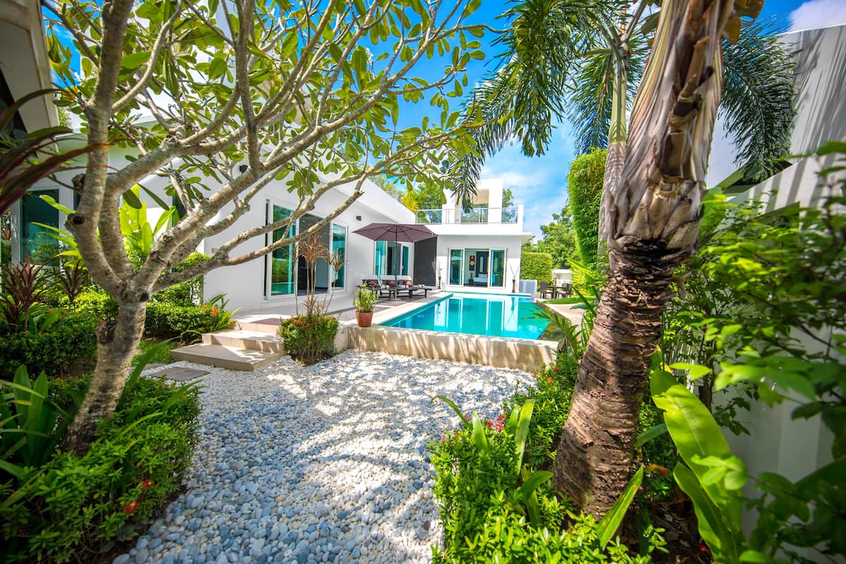 A modern house with a swimming pool, surrounded by lush greenery and a gravel pathway.