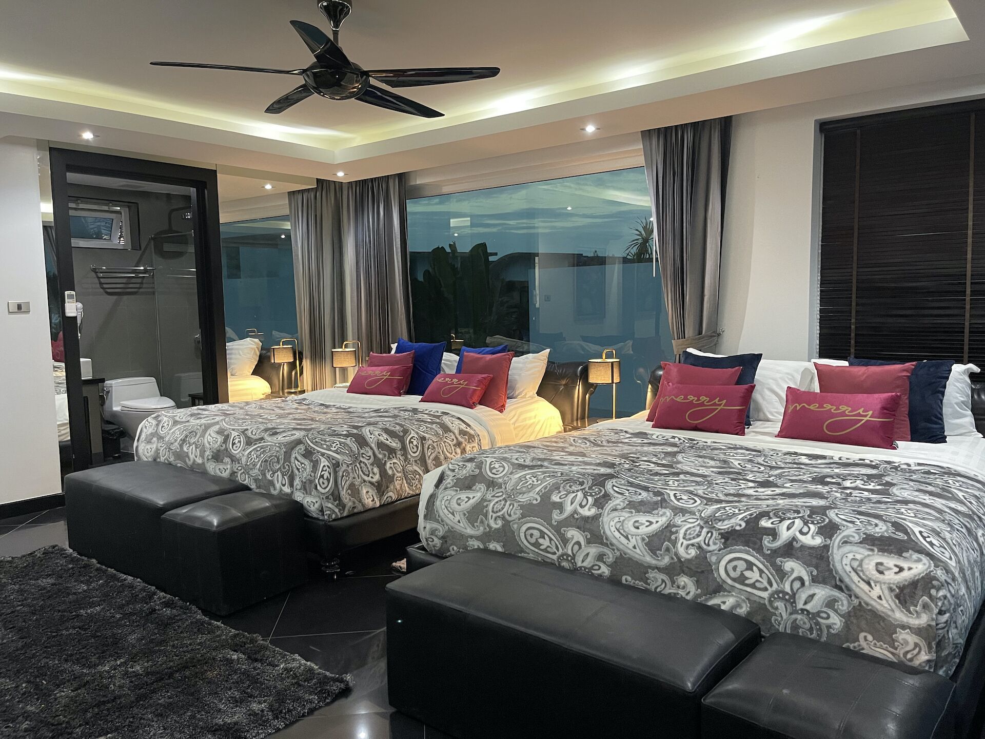 A modern hotel room with two beds, a ceiling fan, large windows, and a view of the outdoors.