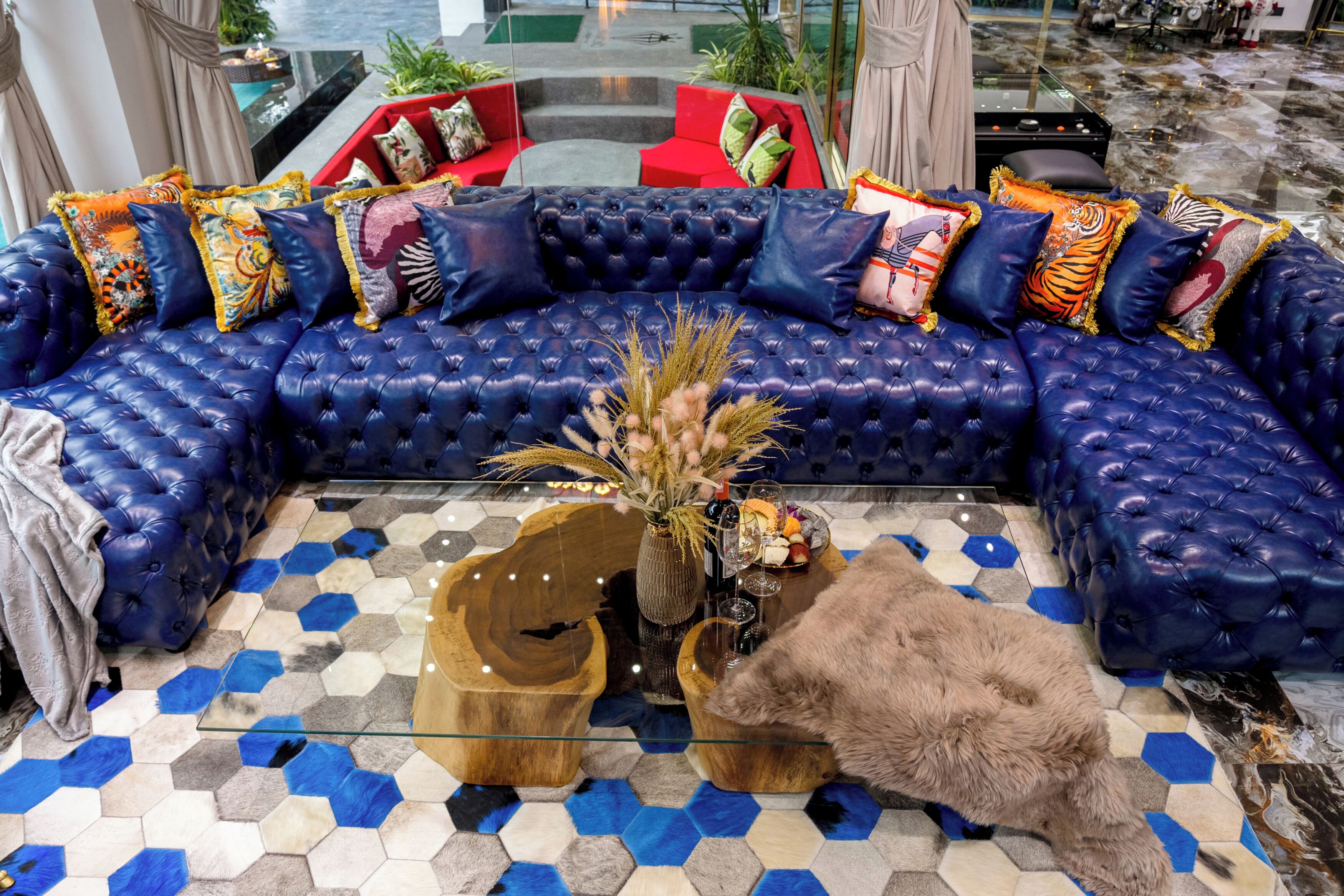 A luxurious blue leather sofa with colorful cushions, a glass coffee table with a vase of dried plants, and a fluffy brown rug.