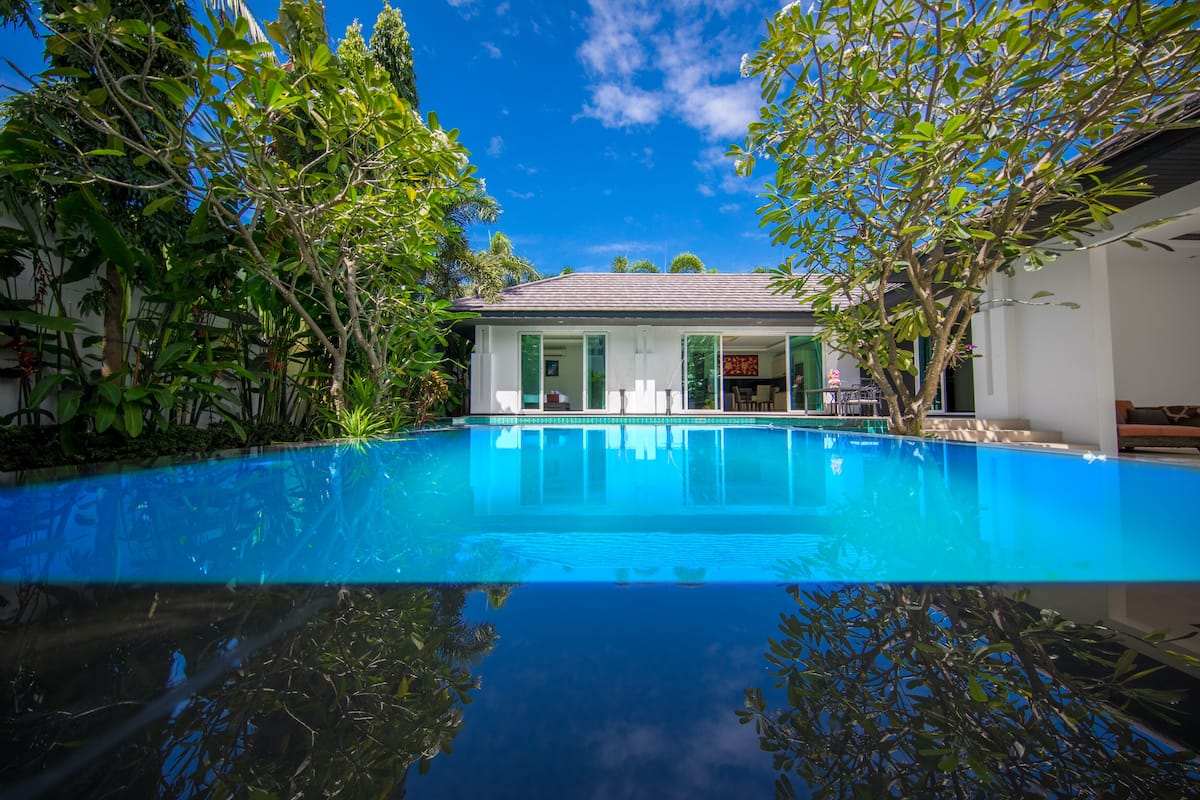 A clear blue swimming pool surrounded by lush greenery and a white building with a tiled roof.