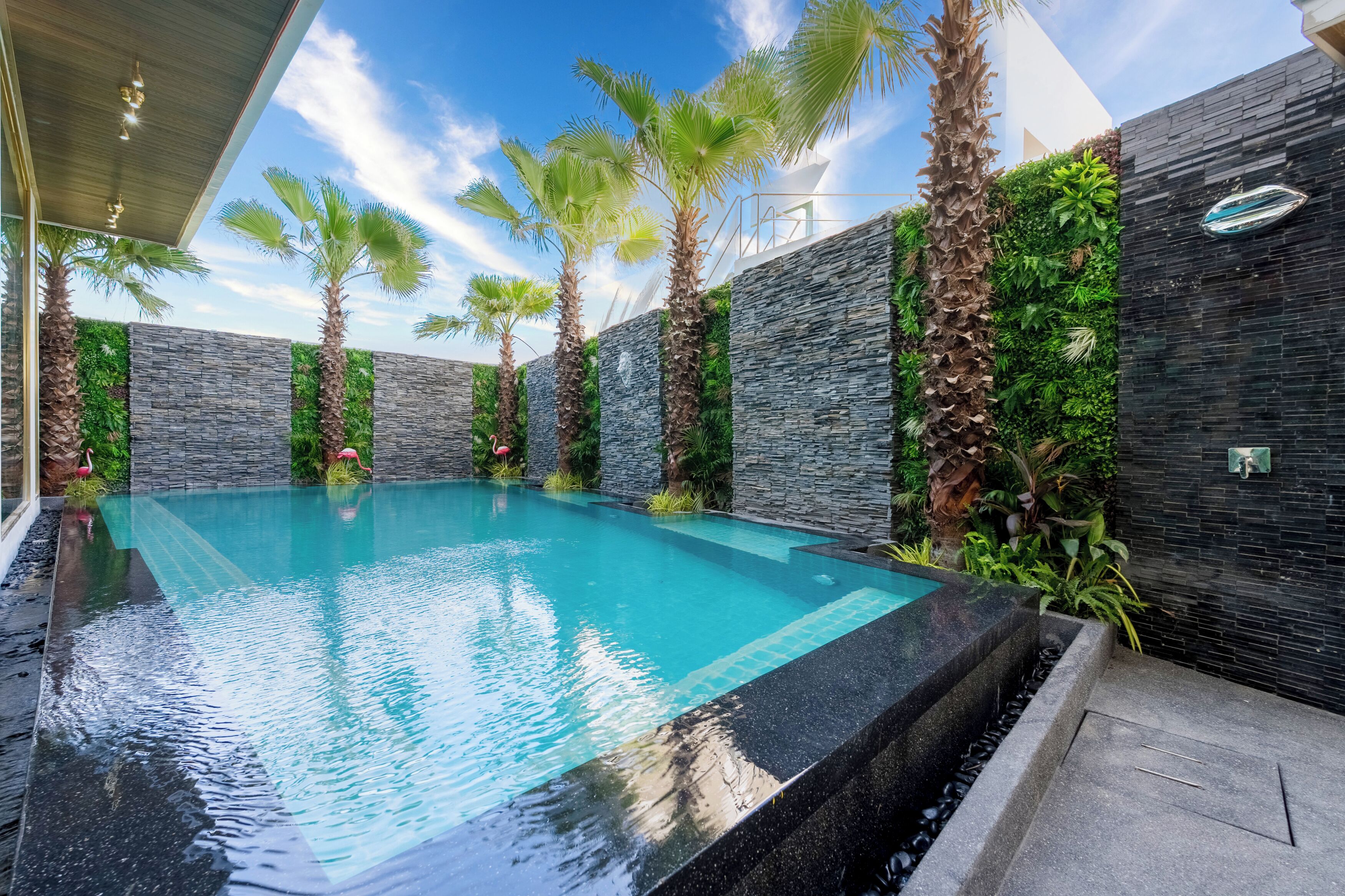 A swimming pool with a stone wall and palm trees, surrounded by a modern building.