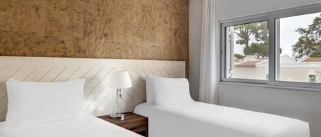 A hotel room with two beds, a wooden headboard, a nightstand with a lamp, and a window with a view of buildings and trees.