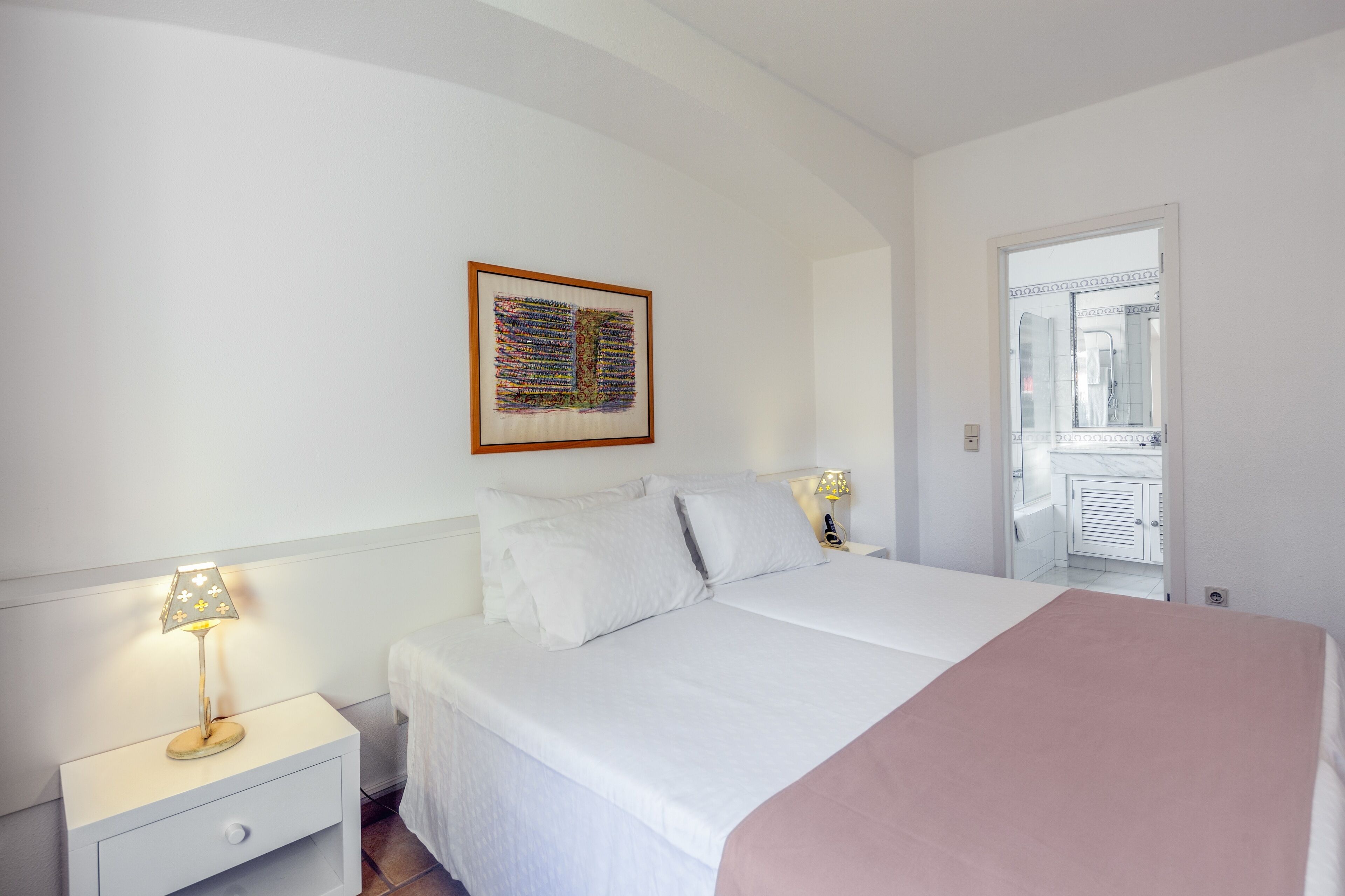 A bedroom with a bed, two bedside tables with lamps, a nightstand, a framed artwork on the wall, and a door leading to a bathroom.