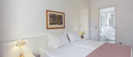 A bedroom with a bed, two bedside tables with lamps, a nightstand, a framed artwork on the wall, and a door leading to a bathroom.