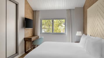 Habitació d'hotel amb llit, escriptori, cadira, televisió i finestra amb vista a arbres.
