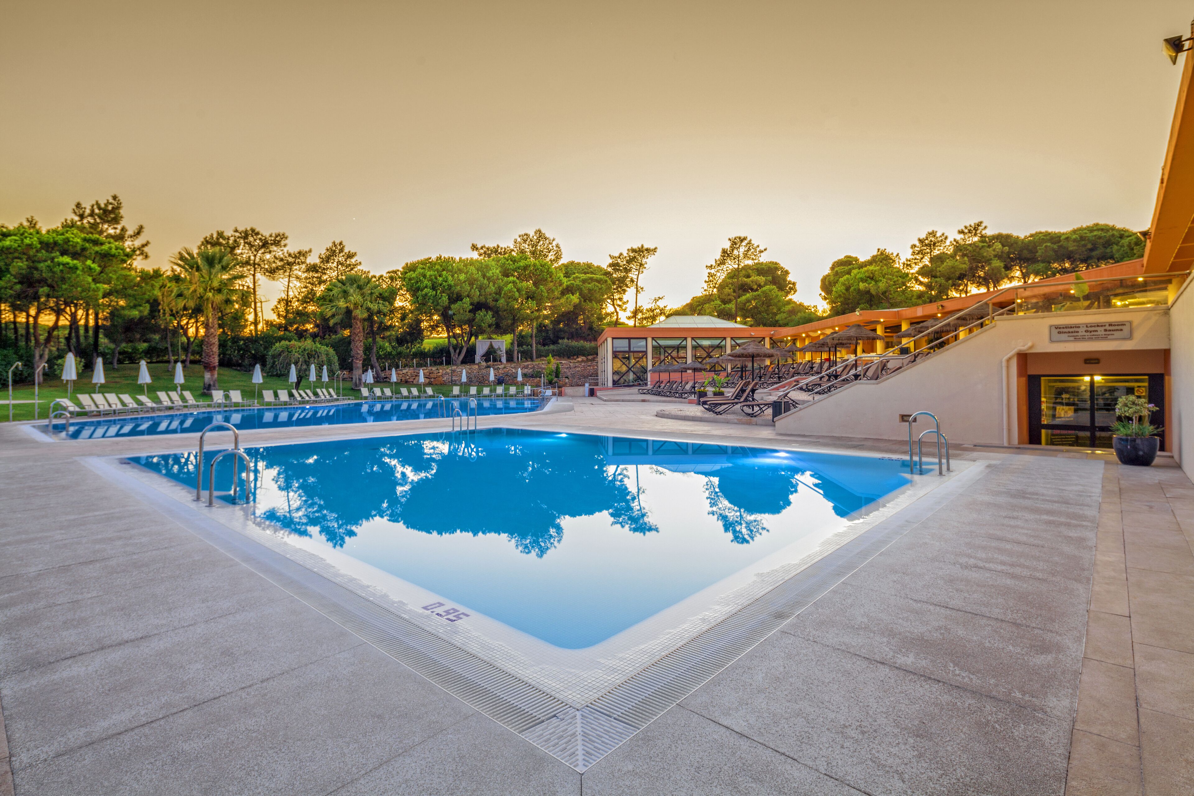 A swimming pool with a clear blue water surface, surrounded by lounge chairs and umbrellas, with a covered seating area and trees in the background.