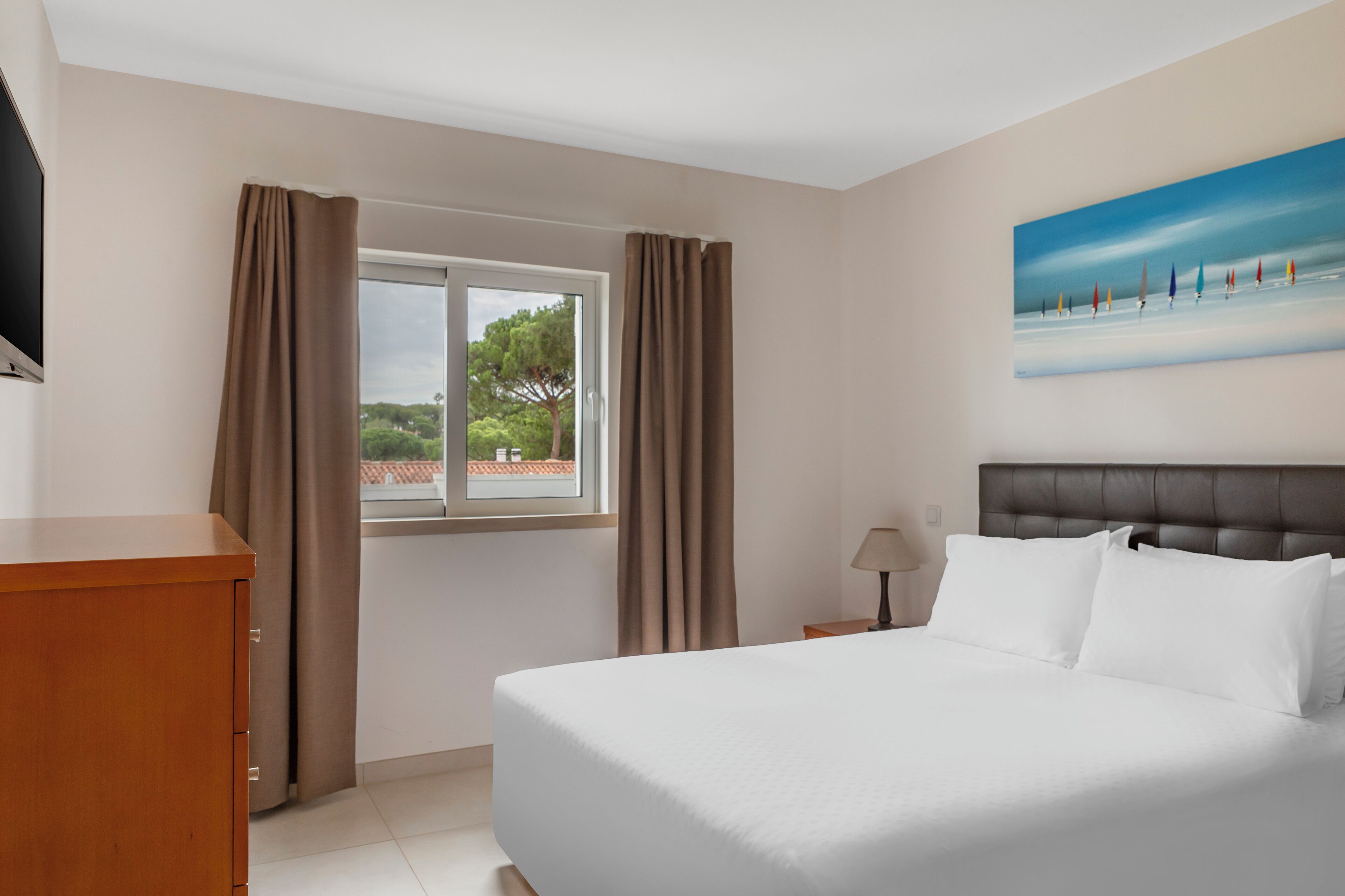 Habitación de hotel con cama, ventana con cortinas, un cuadro en la pared y un aparador de madera.