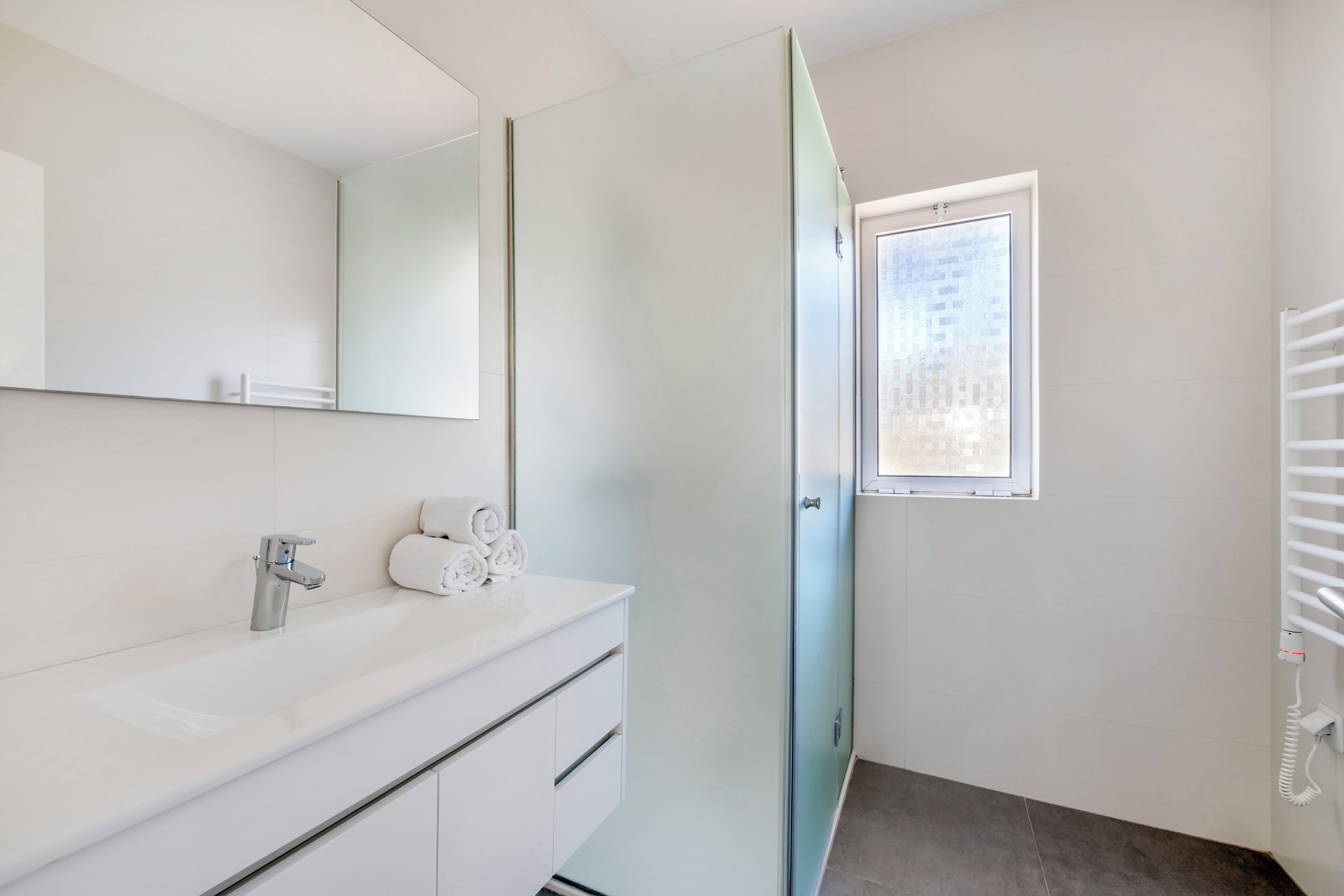 Un baño moderno con tocador blanco, cabina de ducha con mampara de vidrio y ventana que proporciona luz natural.