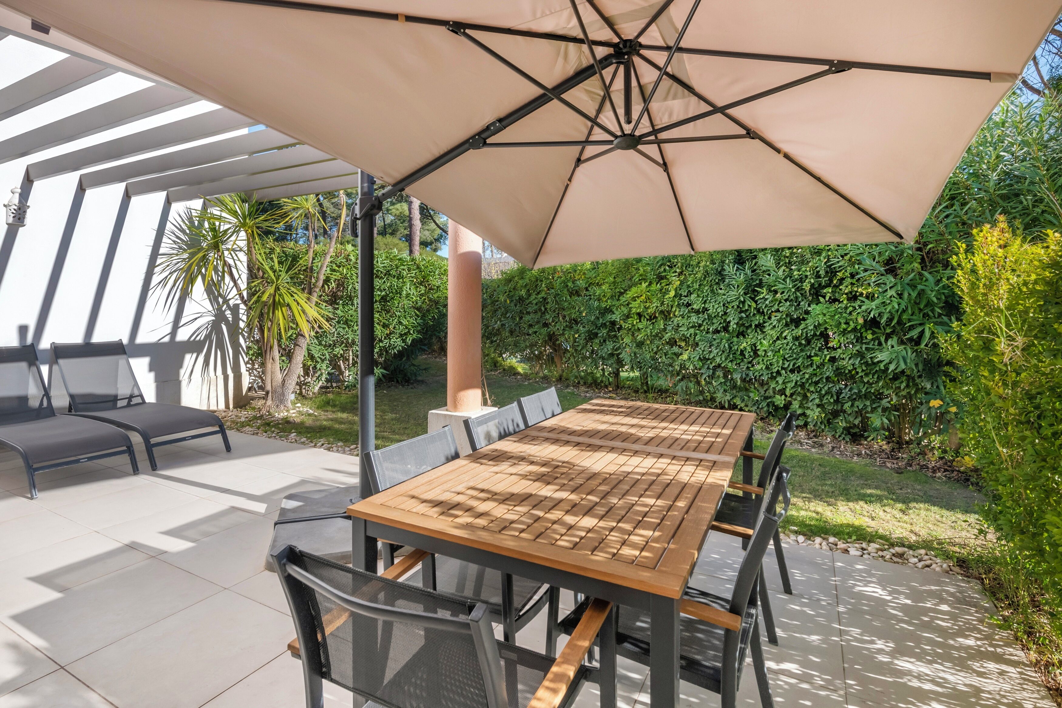 Une terrasse extérieure avec une table et des chaises en bois, un grand parasol et un coin salon agrémenté de coussins.