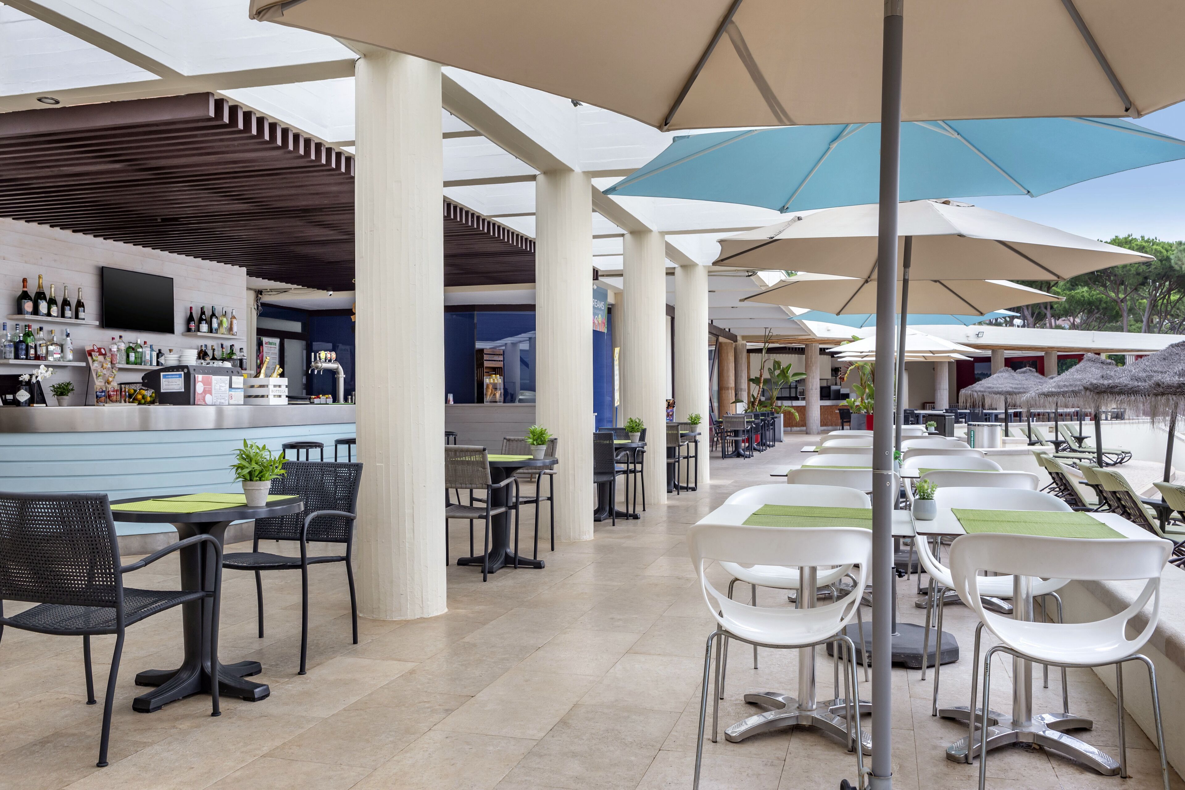 Uma área de café ao ar livre com mesas, cadeiras, guarda-sóis e um bar com garrafas.