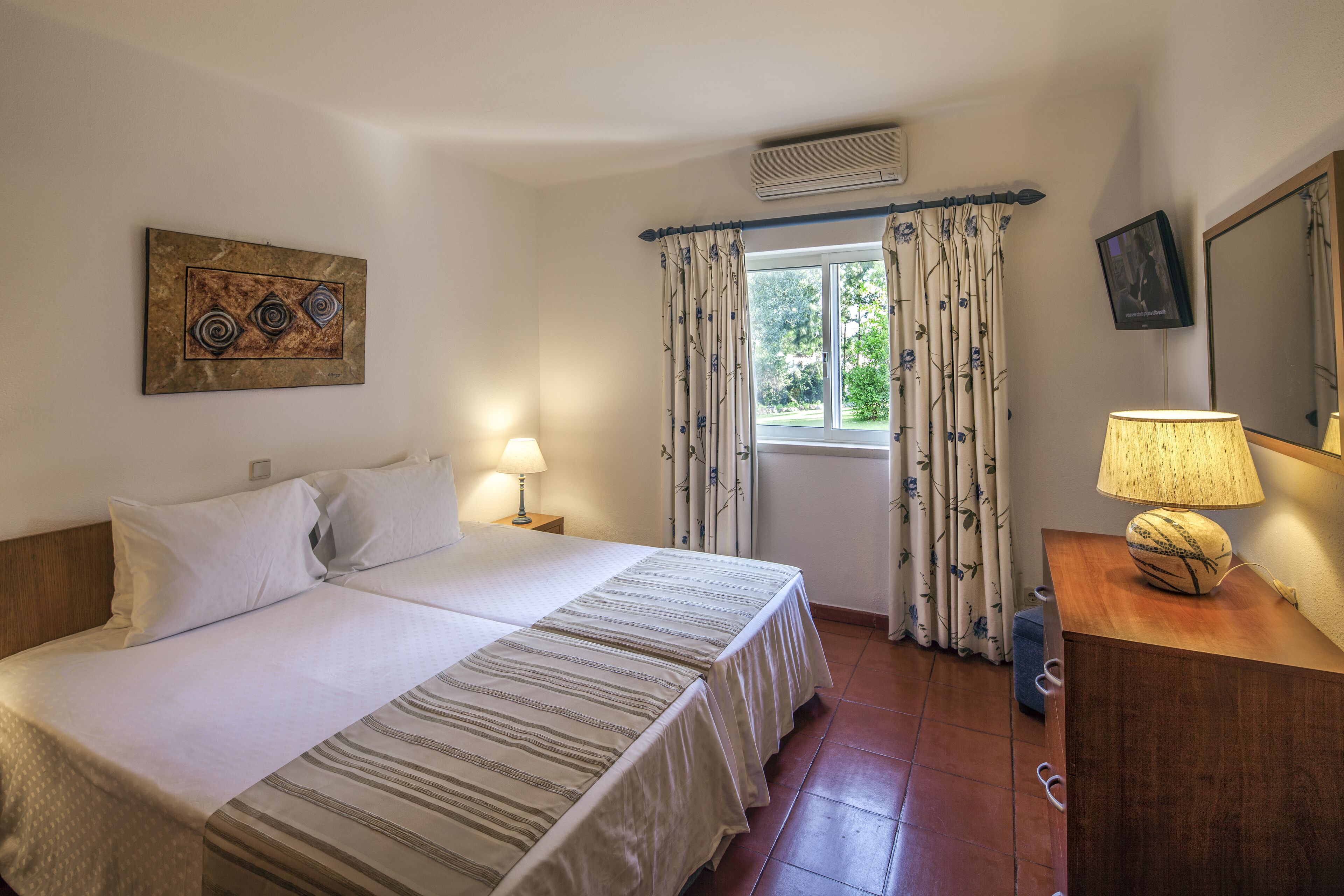 A bedroom with a bed, two bedside tables, a lamp, a mirror, and a window with curtains.