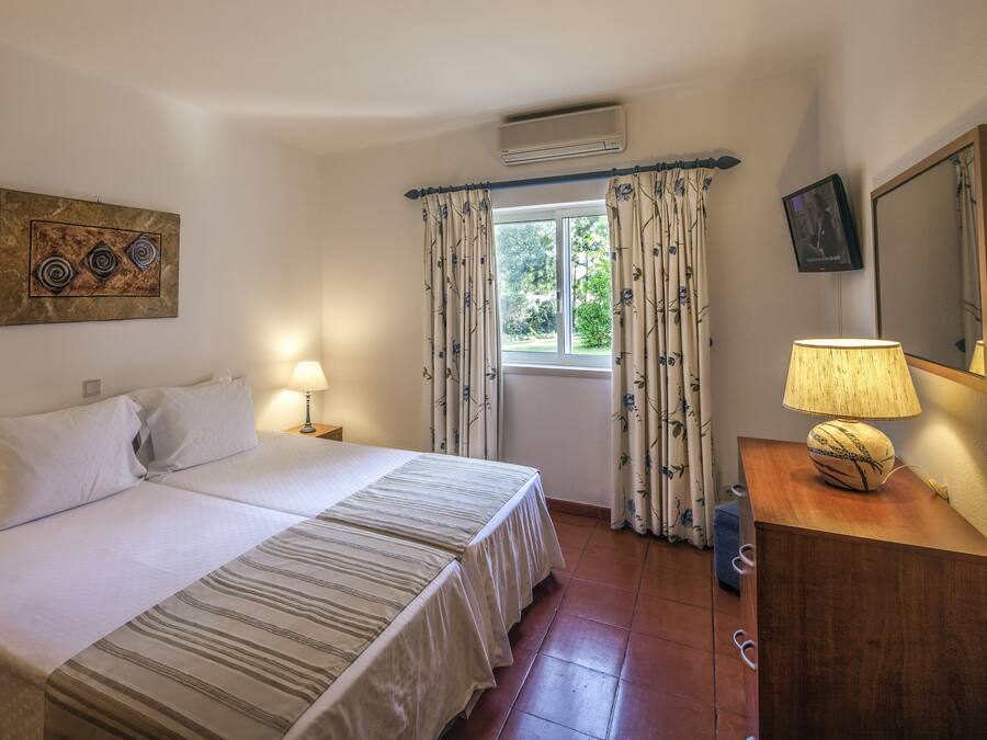 A bedroom with a bed, two bedside tables, a lamp, a mirror, and a window with curtains.