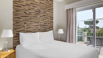 A hotel room with a large bed, a stone wall, a balcony with a view, and two bedside lamps.