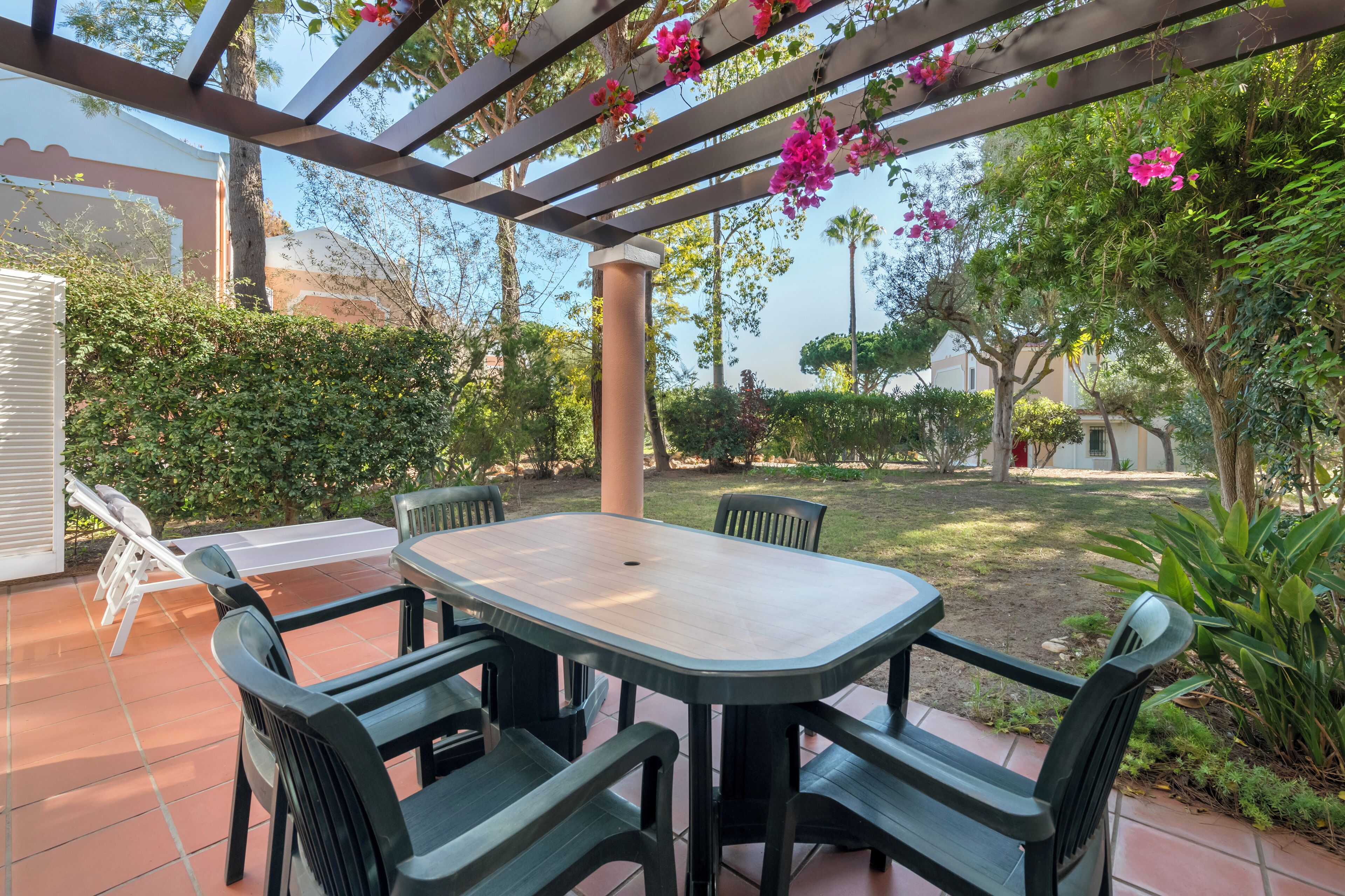 Un patio al aire libre con mesa y sillas, rodeado de follaje y una pérgola adornada con flores.