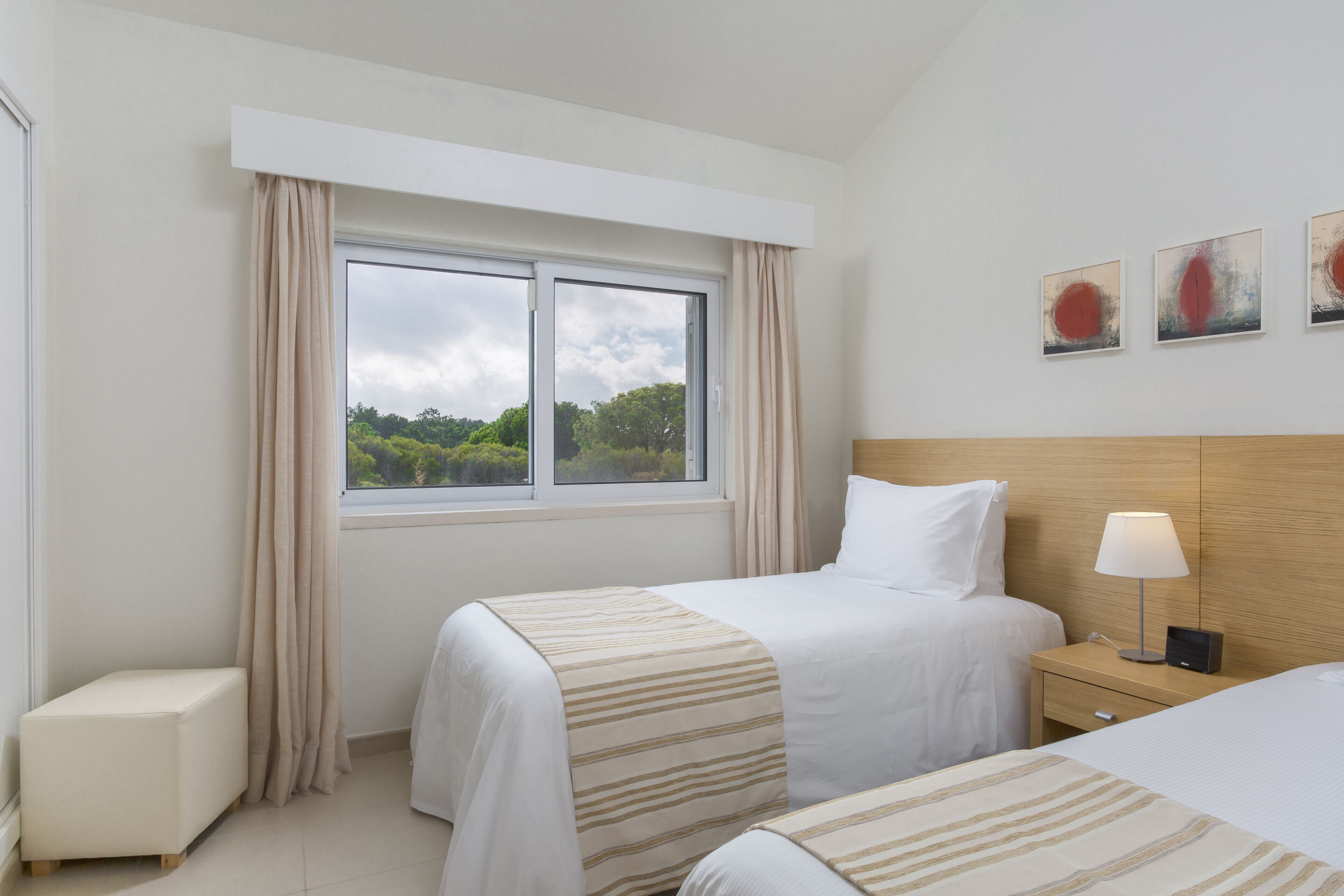 A hotel room with two beds, a bedside table with a lamp, a small bedside table with a phone, a window with curtains, and a view of greenery outside.