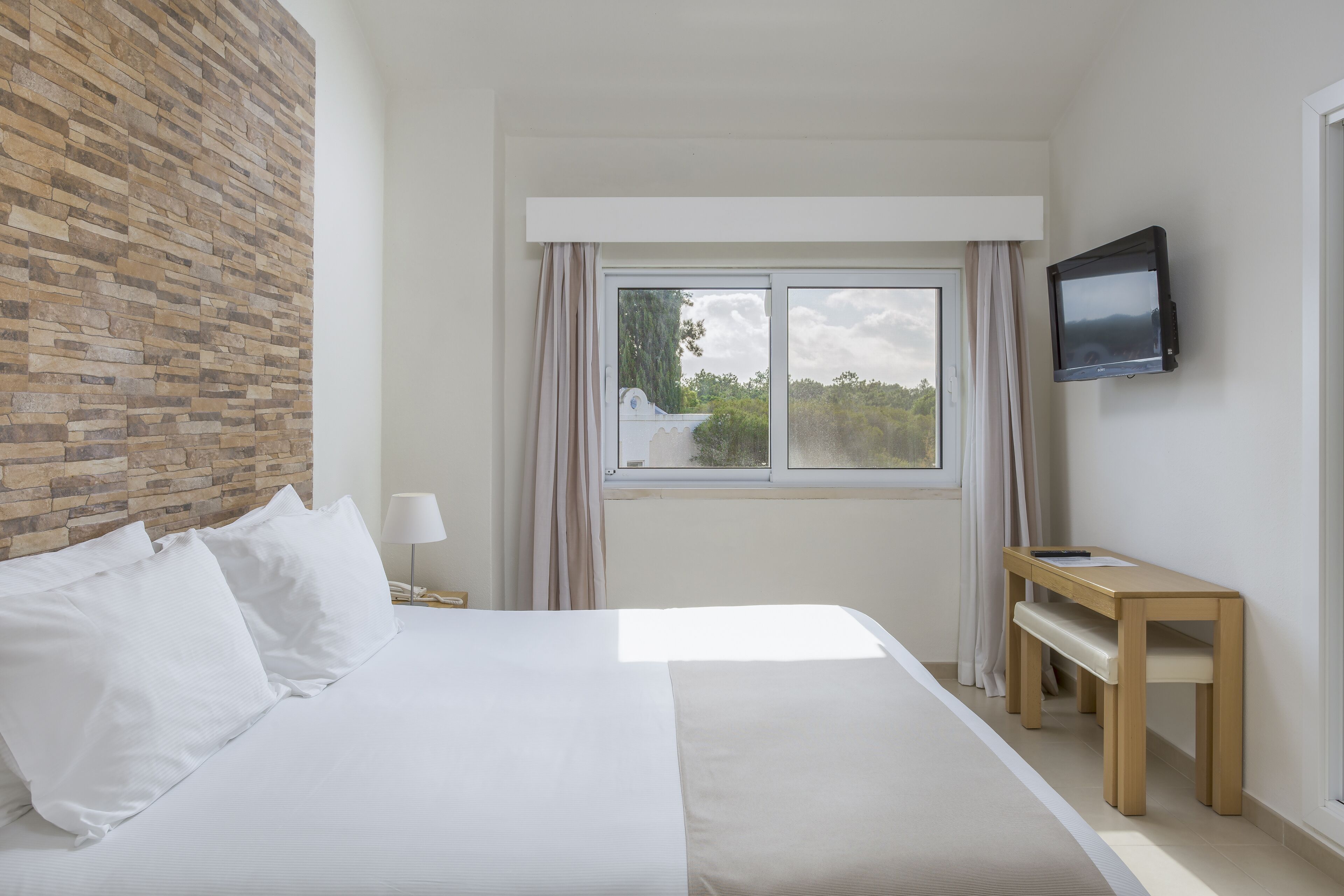 A bedroom with a bed, a wooden nightstand, a TV mounted on the wall, and a window with curtains.