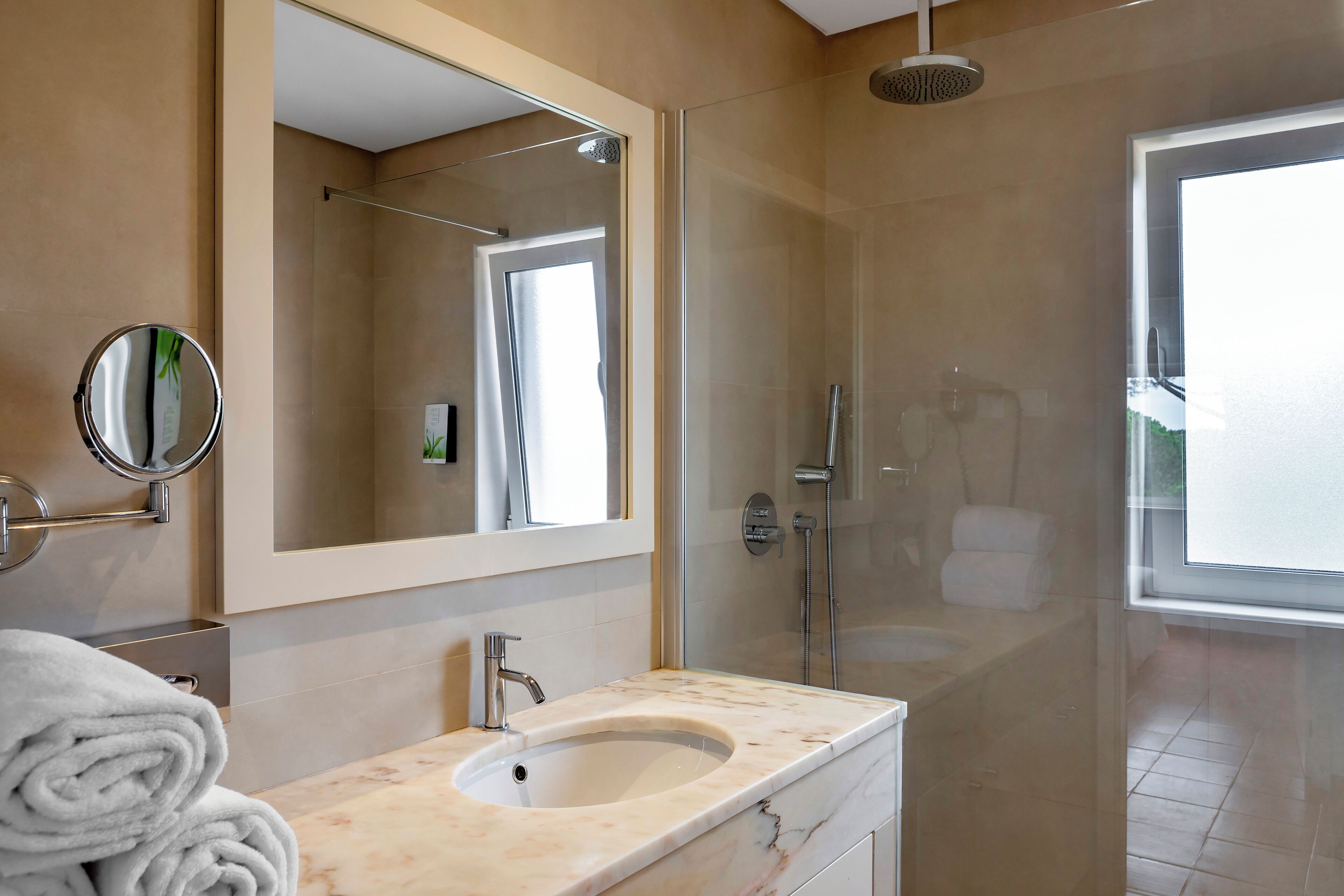 Un baño moderno con lavamanos de mármol, un espejo grande, una ducha con mampara de vidrio y vistas al exterior.