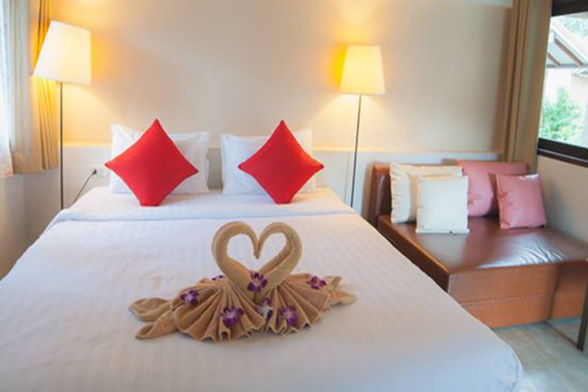 Habitación de hotel con una cama grande, dos almohadas rojas, una disposición de toallas con forma de corazón y una ventana con vistas.