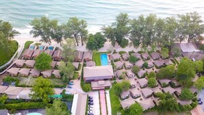 Vista aérea de un complejo turístico con varias cabañas y una piscina central frente a la playa.
