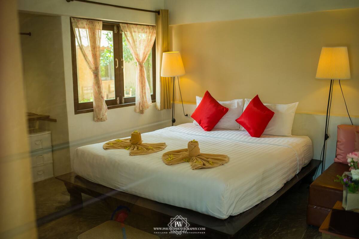 Habitación de hotel con cama, dos lámparas de noche, una ventana con cortinas y una mesita de noche con un jarrón de flores.